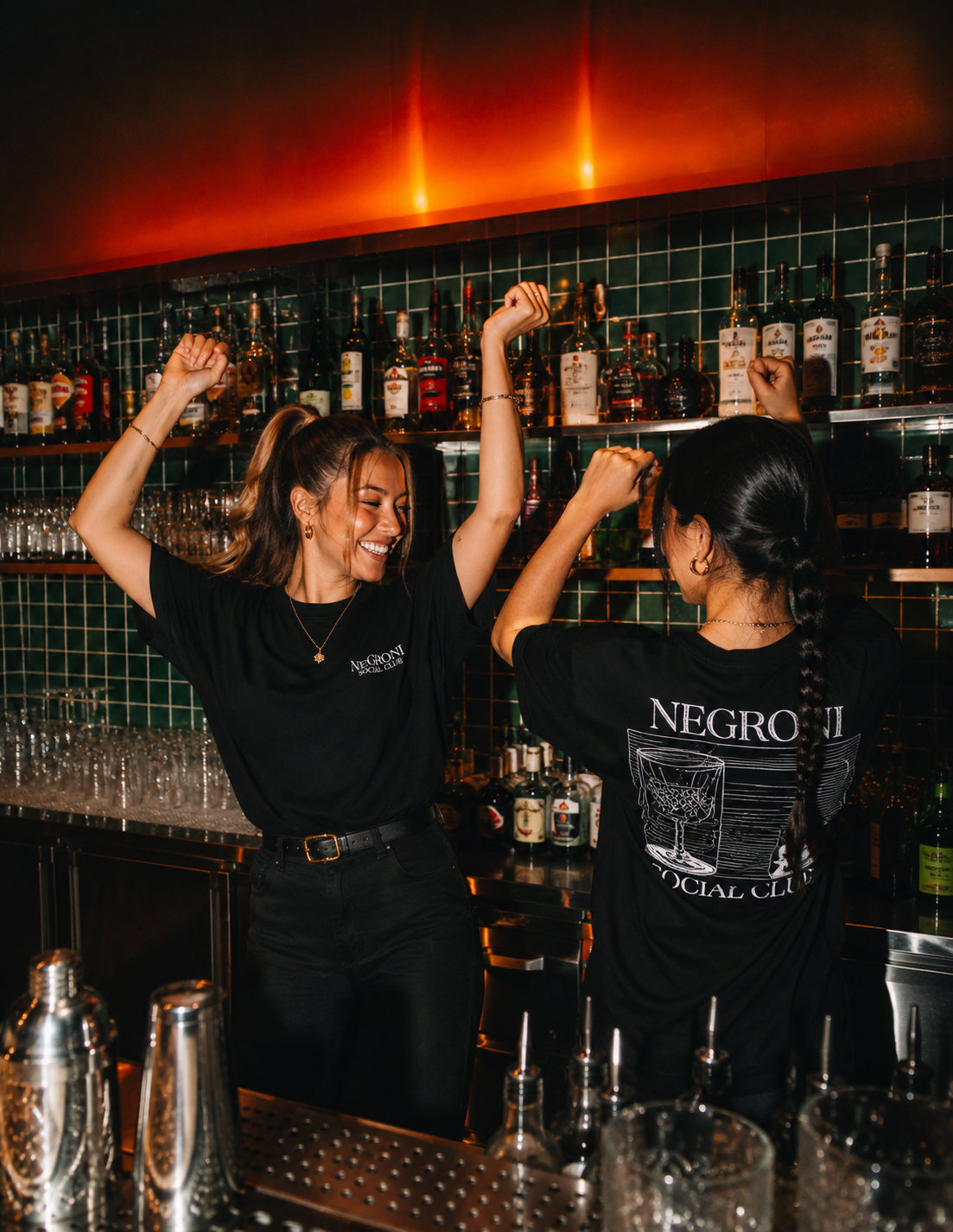 Two women celebrating behind a bar, wearing black T-shirts with 'Negroli Social Club' printed on them, dancing and smiling with arms raised.