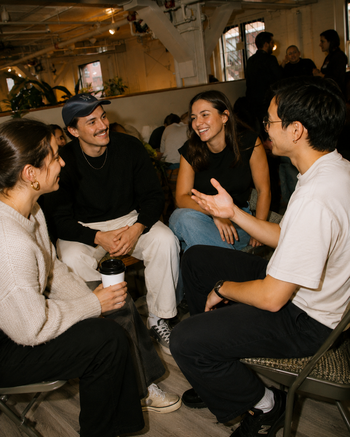 Four young adults sitting in a circle, engaging in conversation, inside a cozy, well-lit cafe or coworking space with large windows and plants in the background.