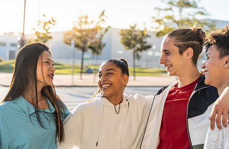 Group of four smiling teenagers standing together outdoors in a park on a sunny day.