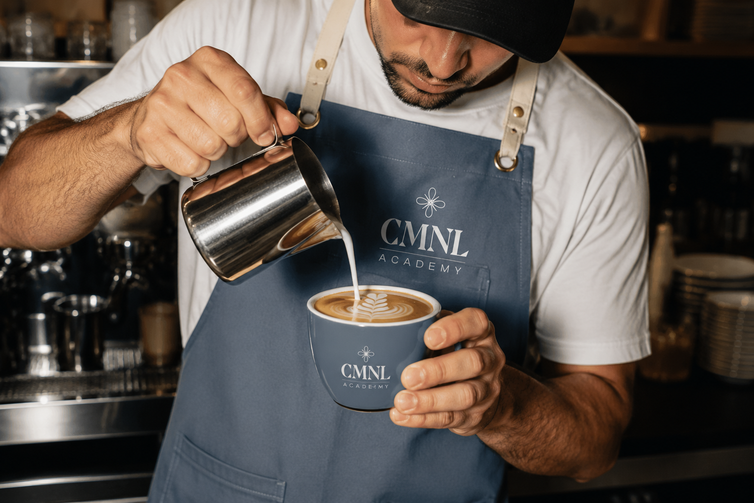 Barista pouring steamed milk into a cup of coffee with latte art, wearing a CMNL Academy apron.