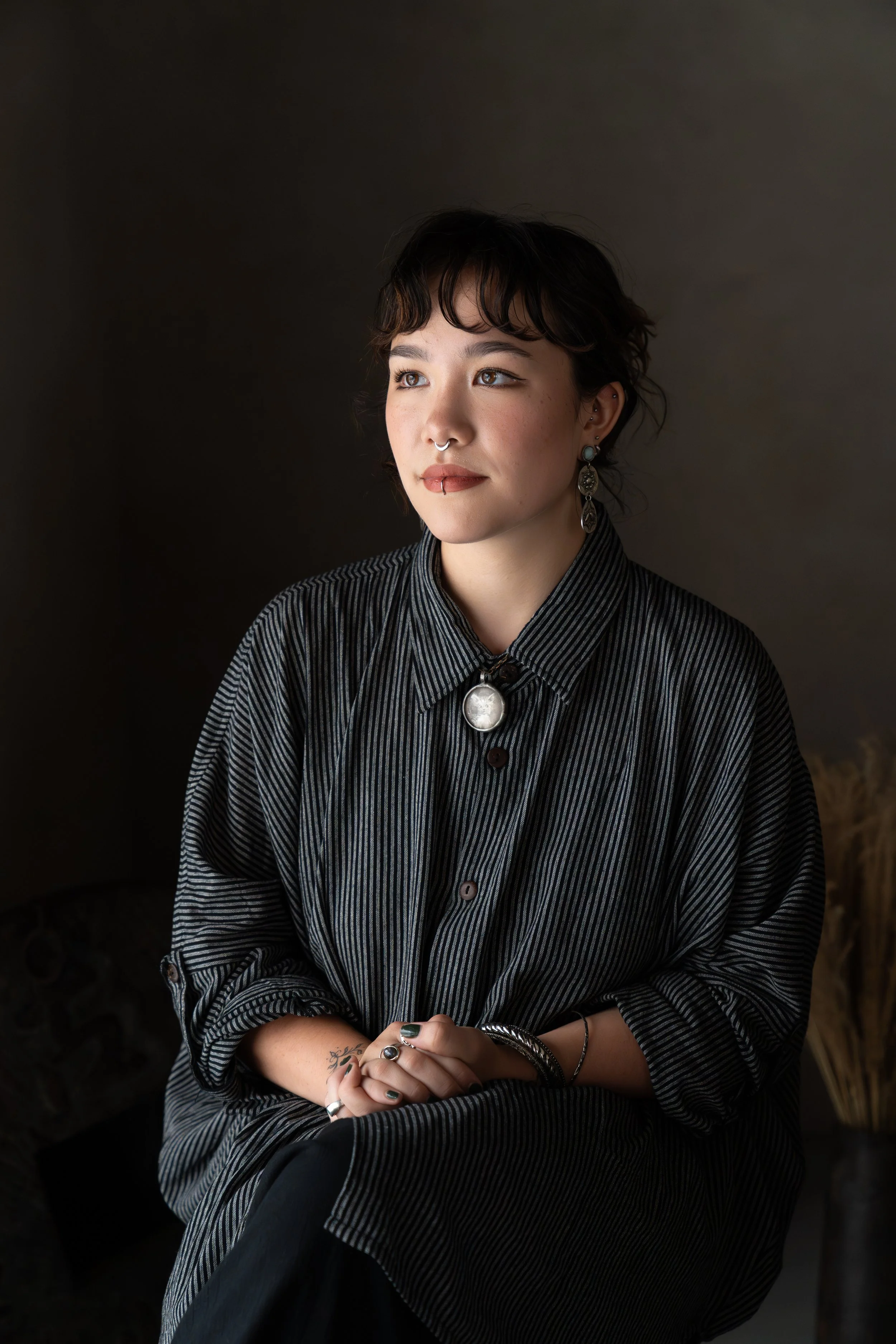 A young woman with short dark hair, wearing a striped shirt and jewelry, sitting against a dark background.