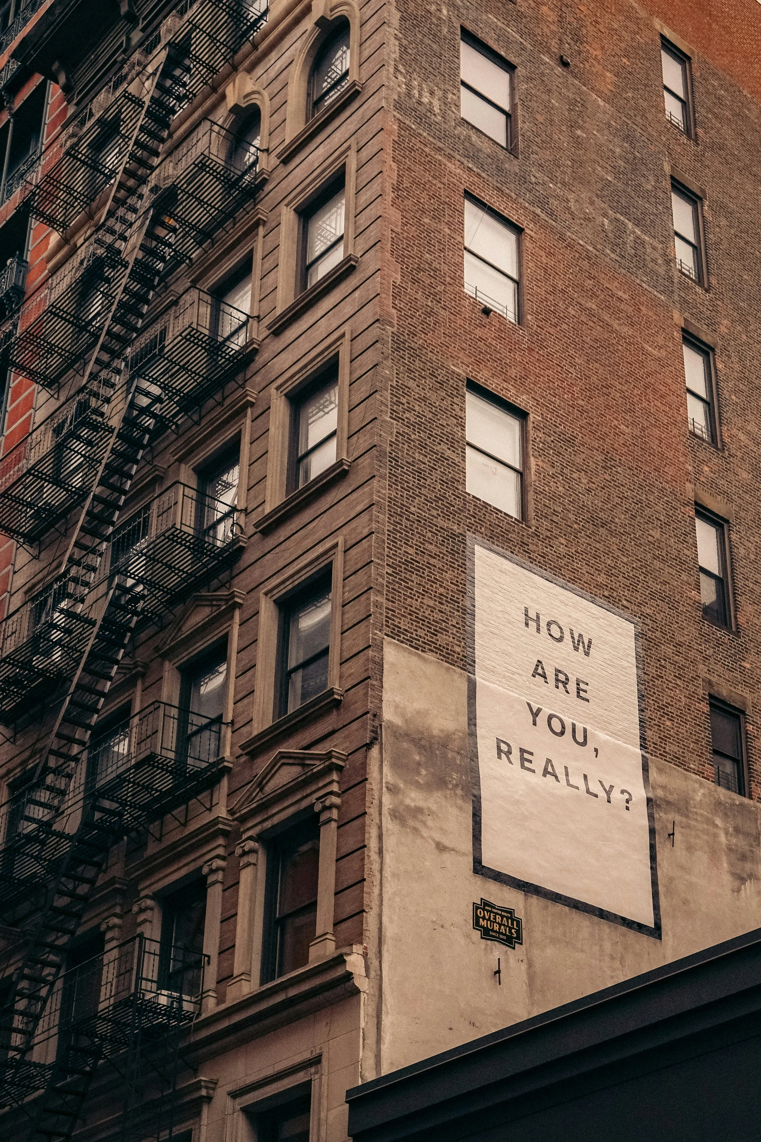 A tall brick building with multiple windows and fire escape stairs on the left side. There is a large white mural on the lower part of the building with the text 'HOW ARE YOU, REALLY?' in black lettering.