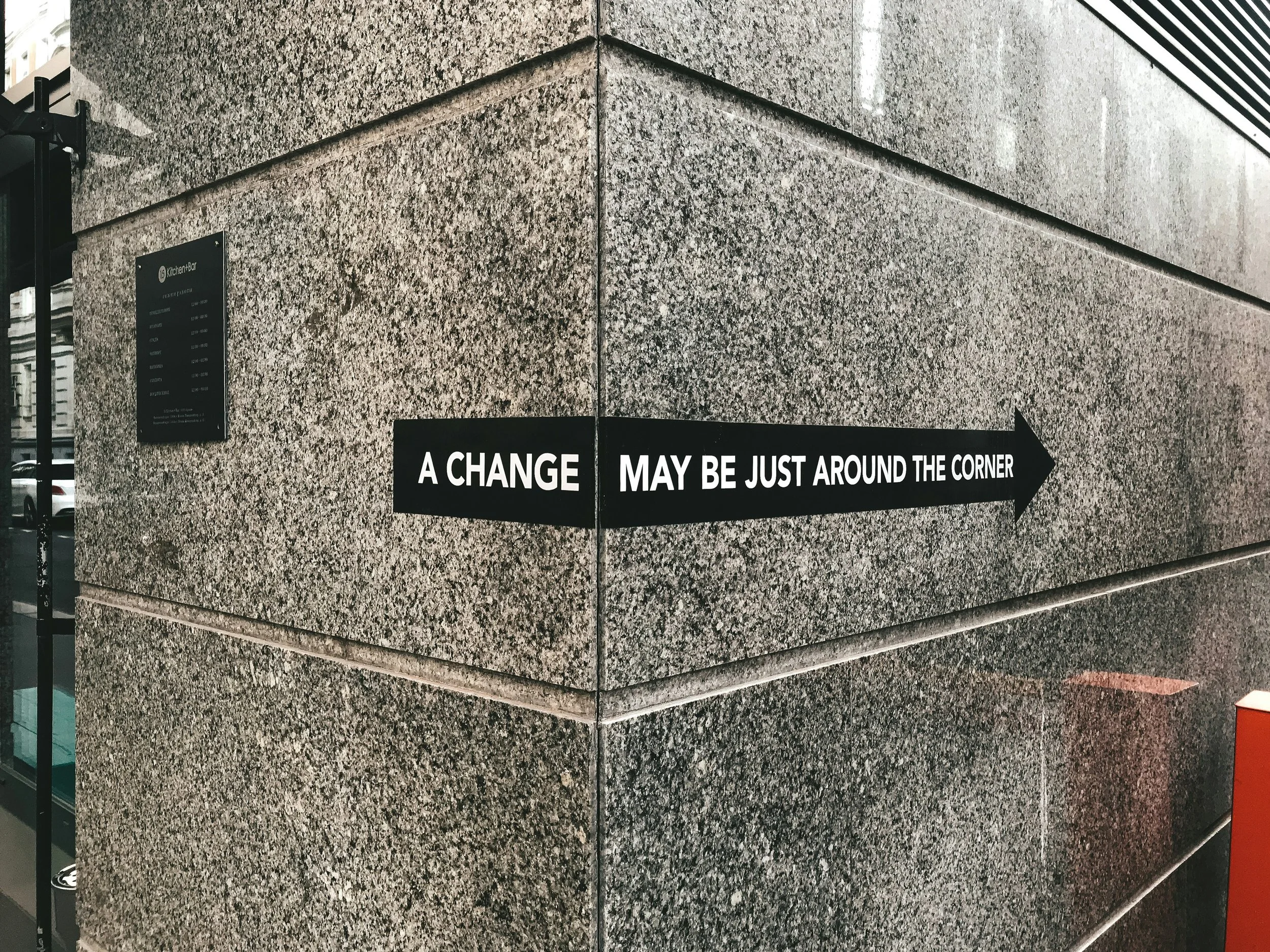 Black arrow-shaped sign on a beige granite wall with the text 'A CHANGE MAY BE JUST AROUND THE CORNER'.