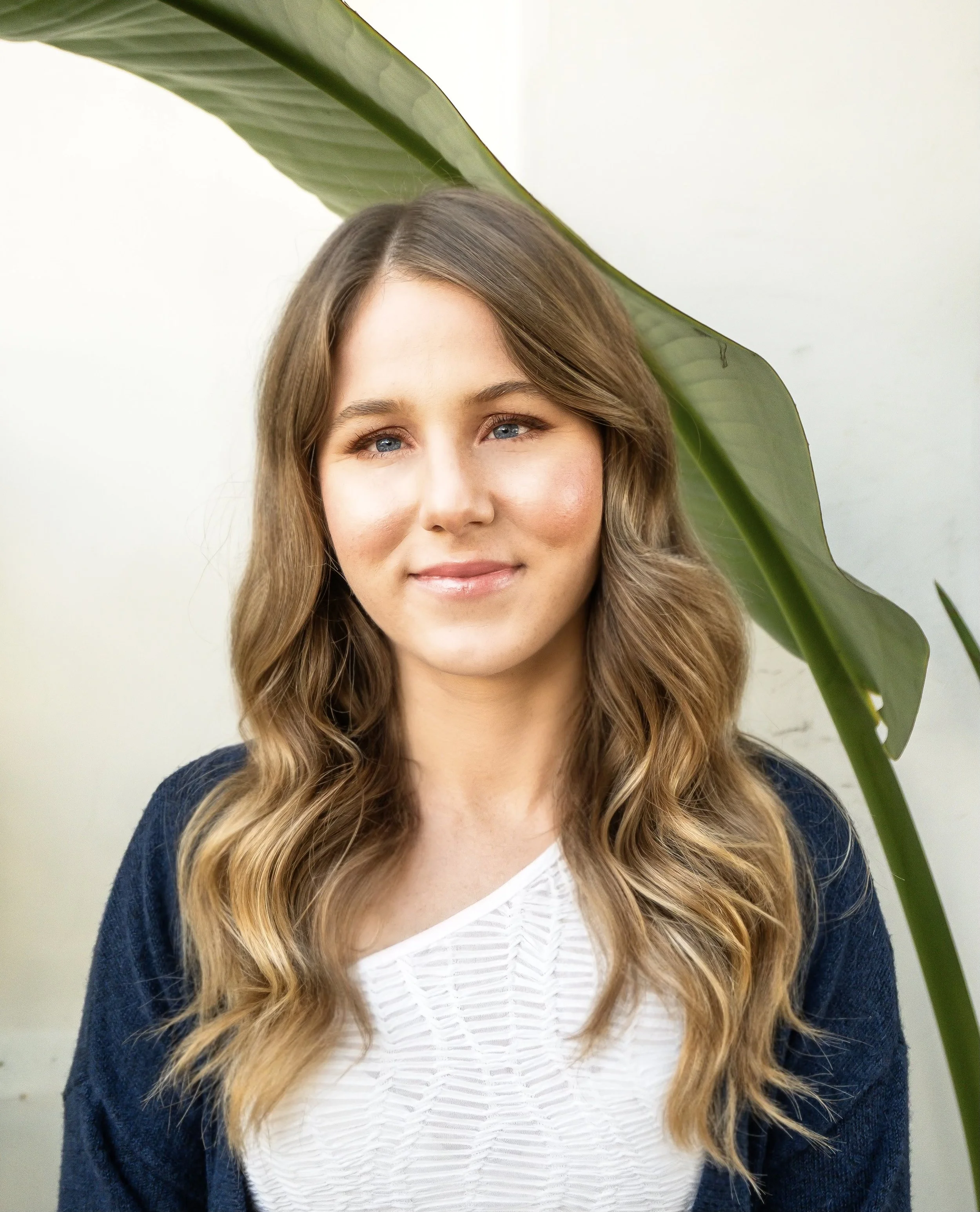 A young woman with wavy, light brown hair and blue eyes, smiling softly, wearing a white top and navy blazer, standing in front of a white wall with large green leaves partially covering her.