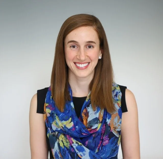 Young woman with shoulder-length red hair, wearing a sleeveless black top and a colorful blue, purple, and yellow patterned scarf, smiling against a plain gray background.
