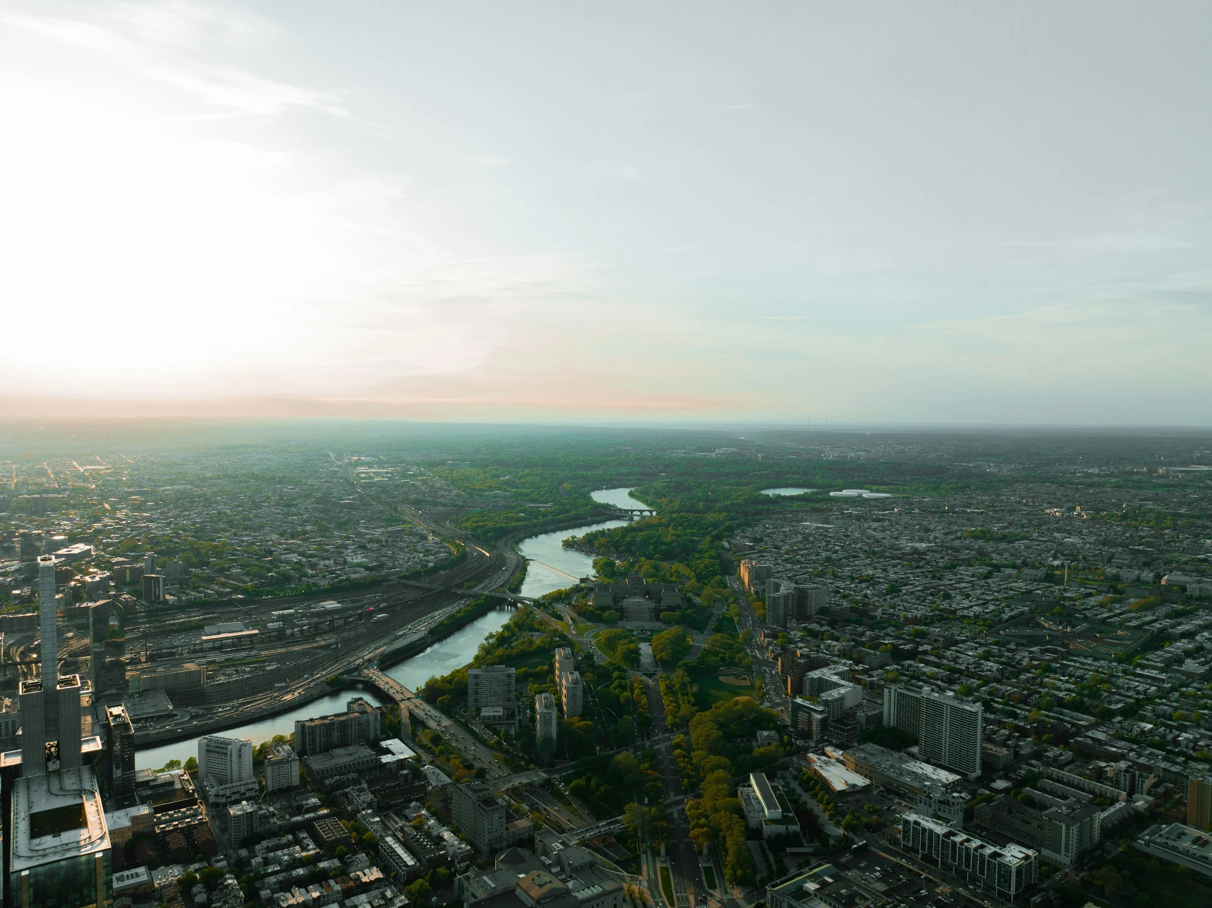 Aerial view of a cityscape with a winding river, green parks, and numerous buildings at sunset or sunrise.