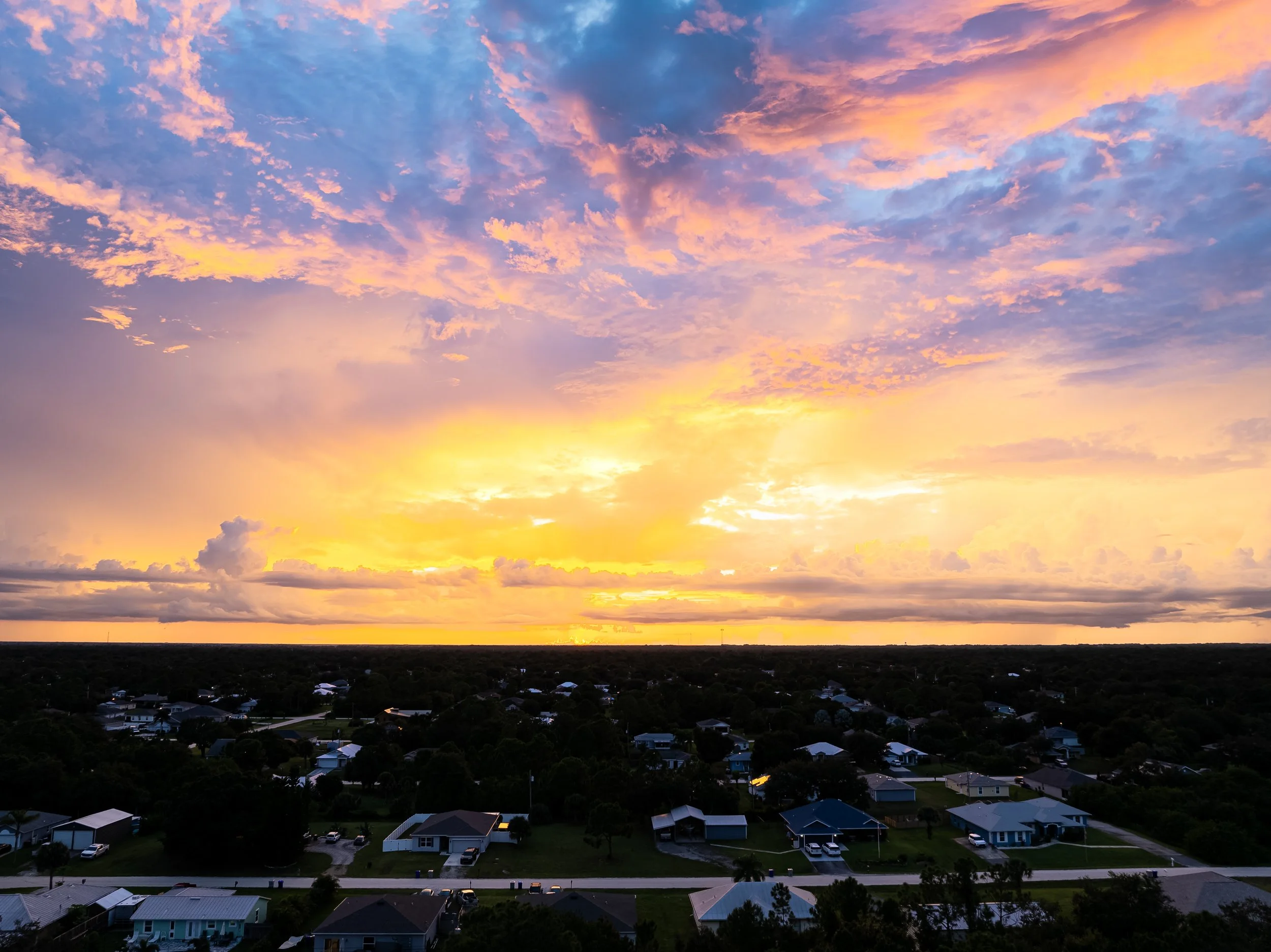 A scenic view of a sunset over a suburban neighborhood with houses and trees, under a vibrant sky with pink, purple, and yellow clouds.