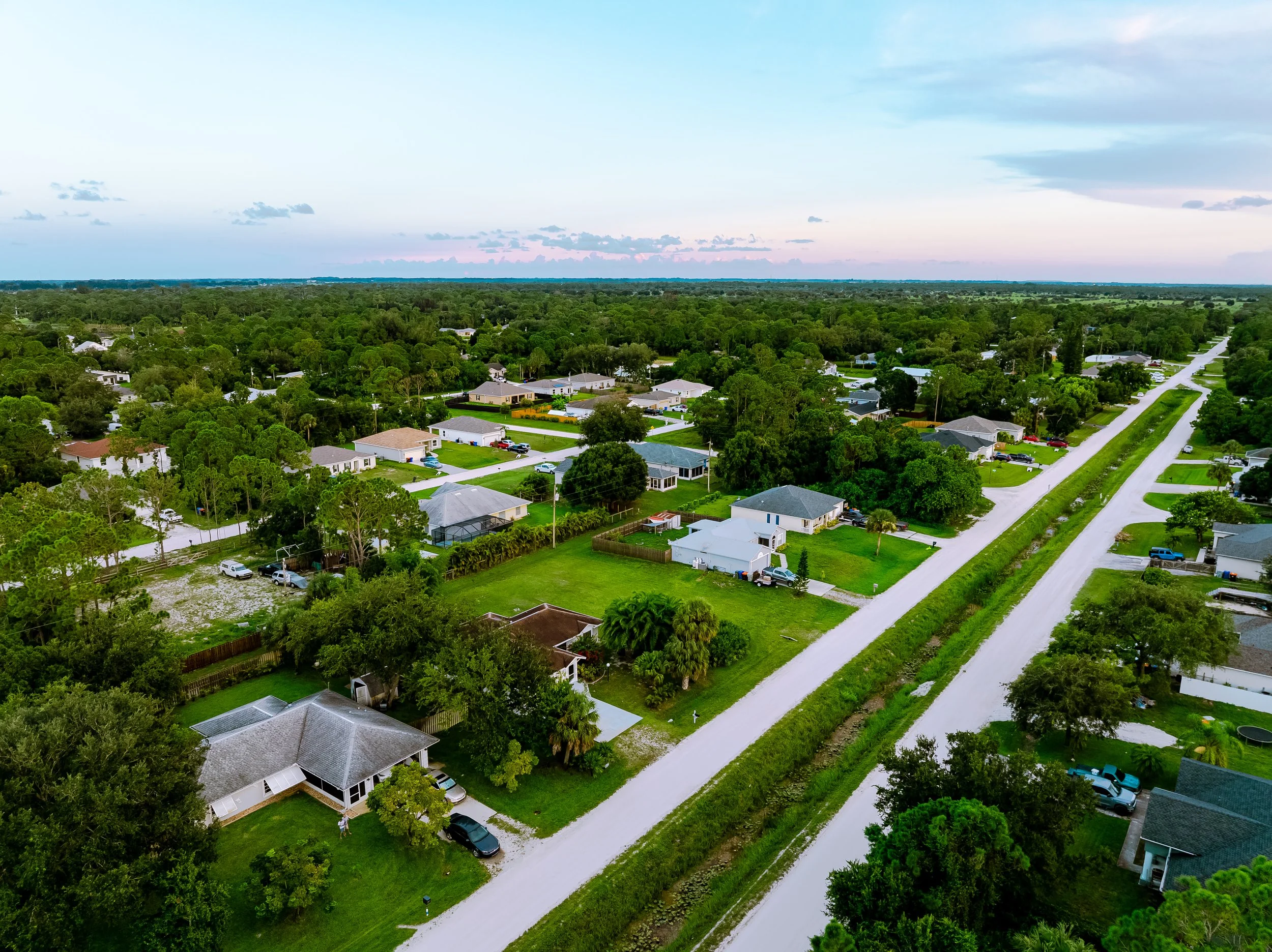 An aerial view of a suburban neighborhood shows houses with yards, trees, and parked cars on a dirt road during daytime with a partly cloudy sky.