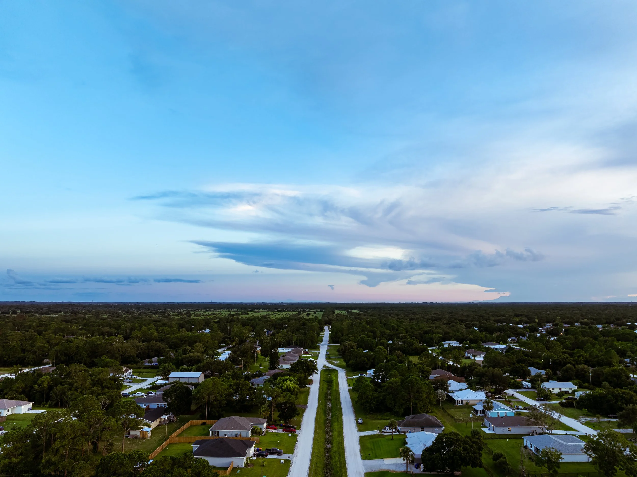 Aerial view of a suburban neighborhood with single-family homes, green lawns, and trees, extending toward the horizon under a partly cloudy sky during sunset.