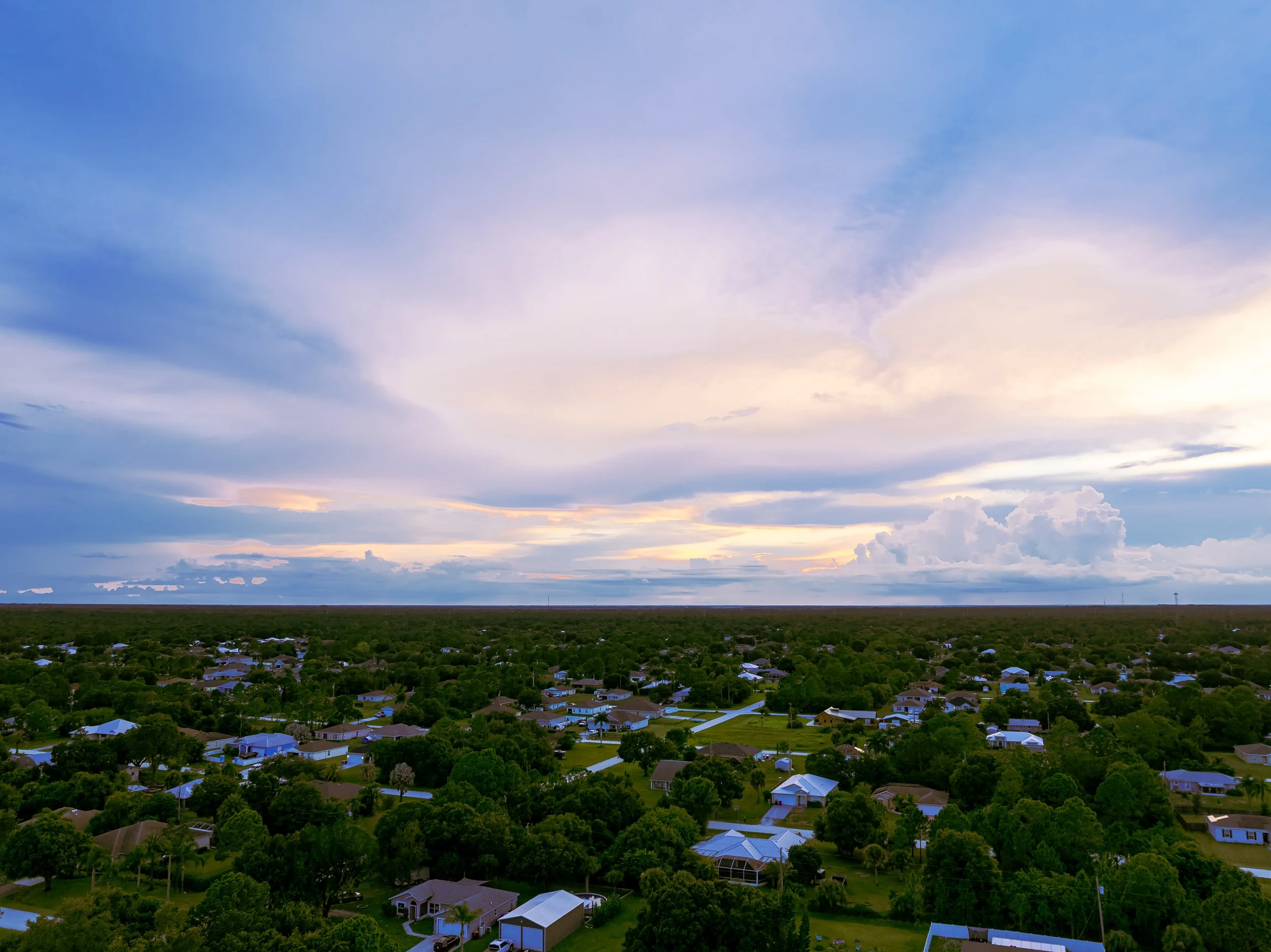 An aerial view of a suburban neighborhood at sunset with houses surrounded by trees and a large sky with clouds.