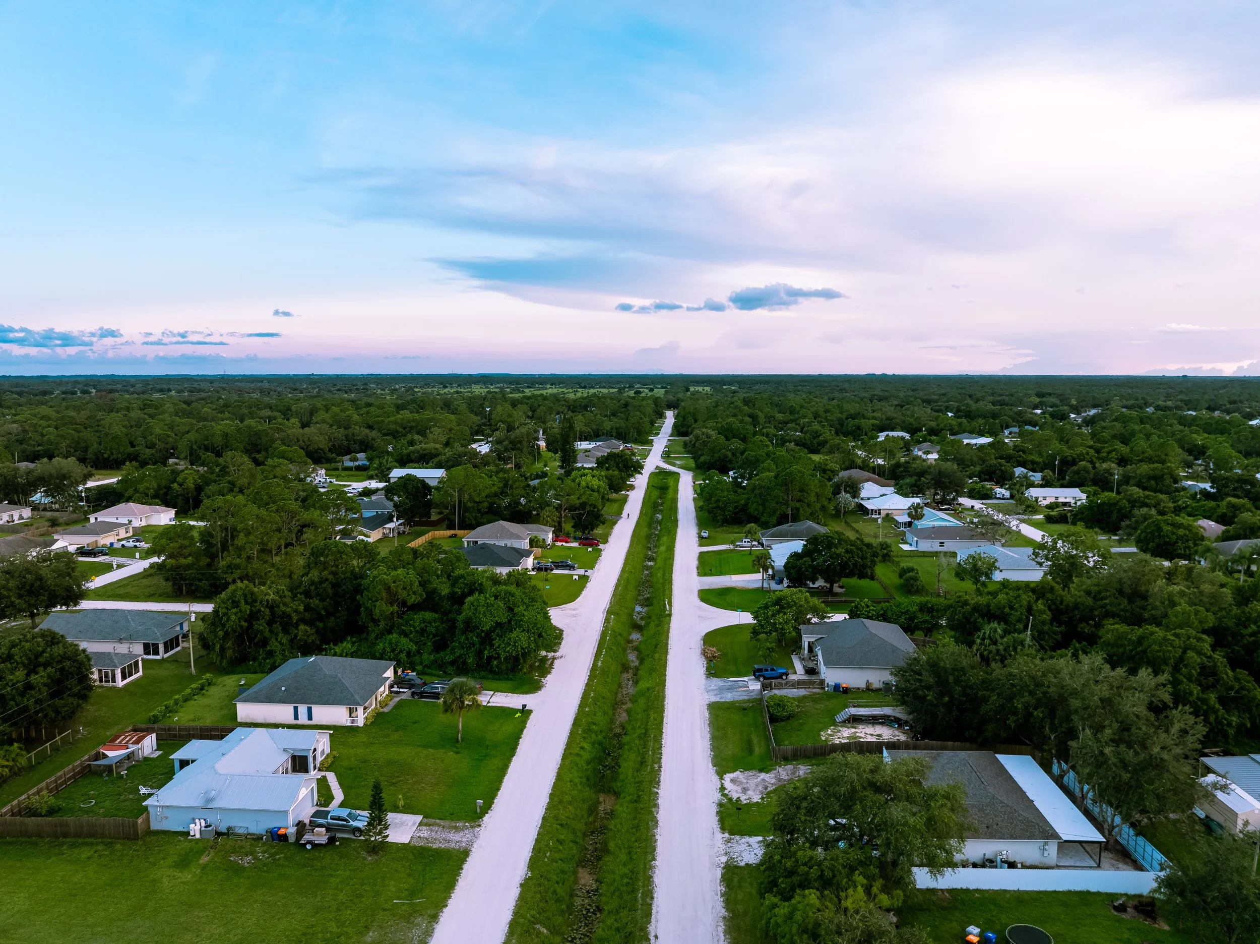 An aerial view of a suburban neighborhood with houses on both sides of a dirt road, surrounded by green trees and open land, under a partly cloudy sky.