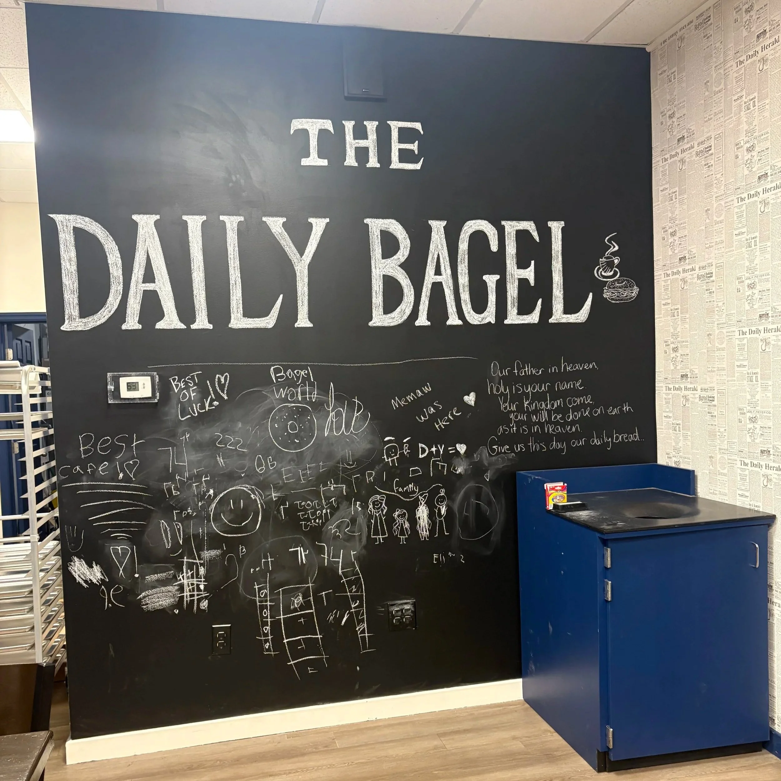 Black chalkboard wall with white chalk writing entitled "The Daily Bagel," featuring drawings, messages, and doodles, including a smiley face, family stick figures, and references to coffee and good luck, with a blue cabinet beside it.