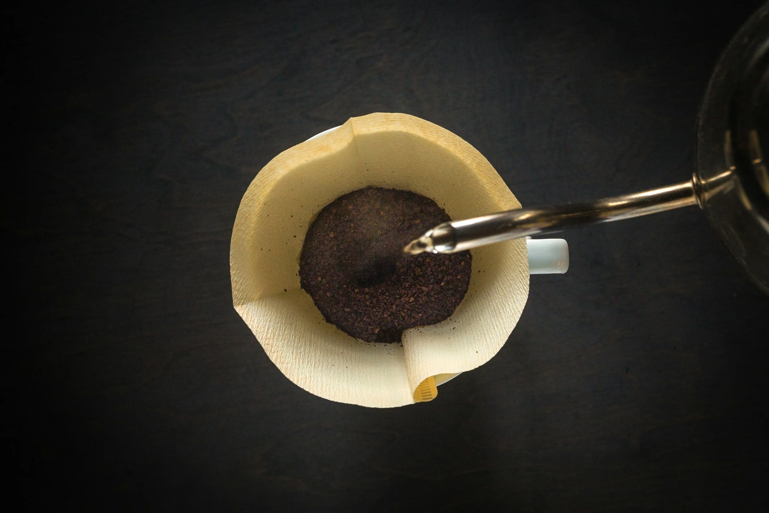 A coffee filter with ground coffee, with a gooseneck kettle pouring hot water into it, atop a dark surface.