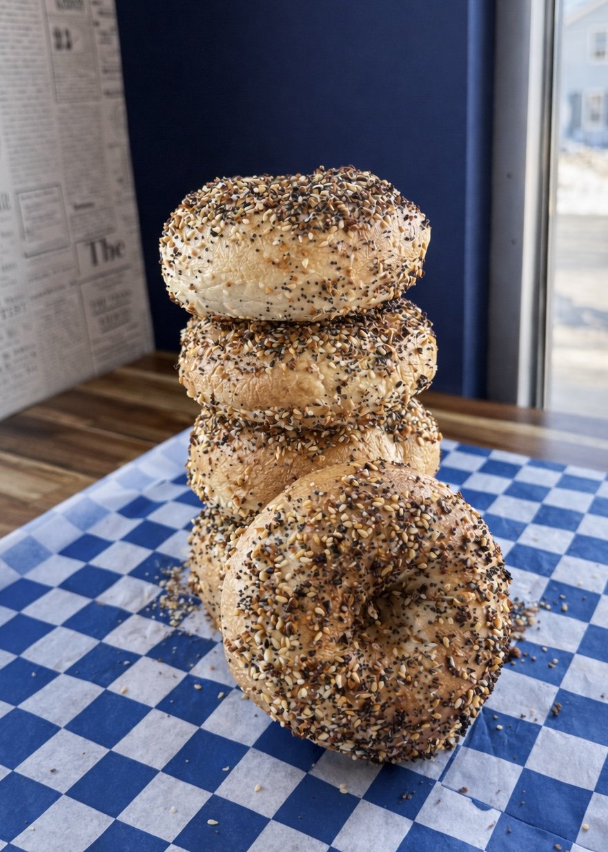 Four sprinkled bagels stacked on top of each other on a checkered paper surface, with a window and wall in the background.