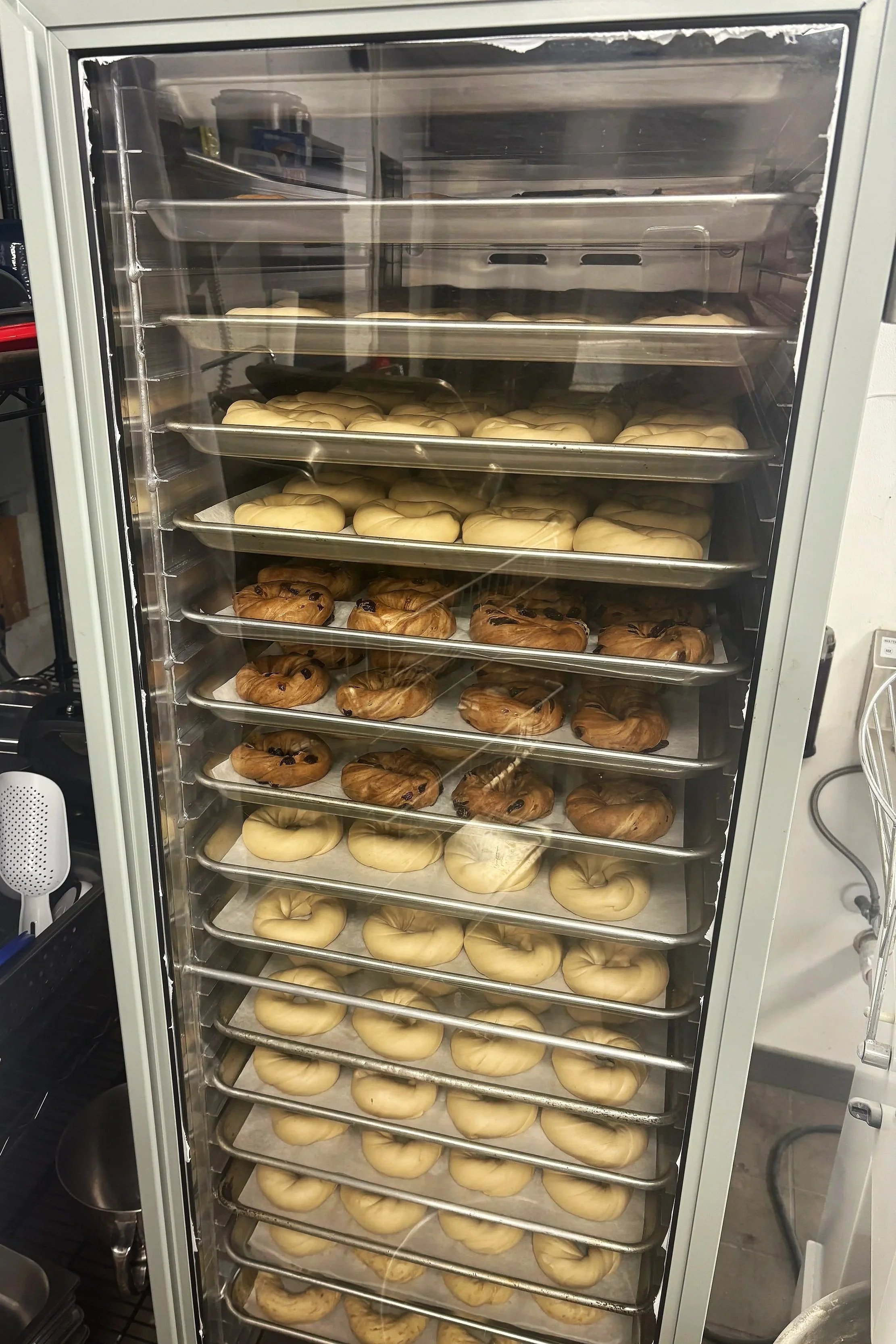 A commercial oven with multiple racks filled with raw dough and baked cookies, including chocolate chip cookies and bagels.