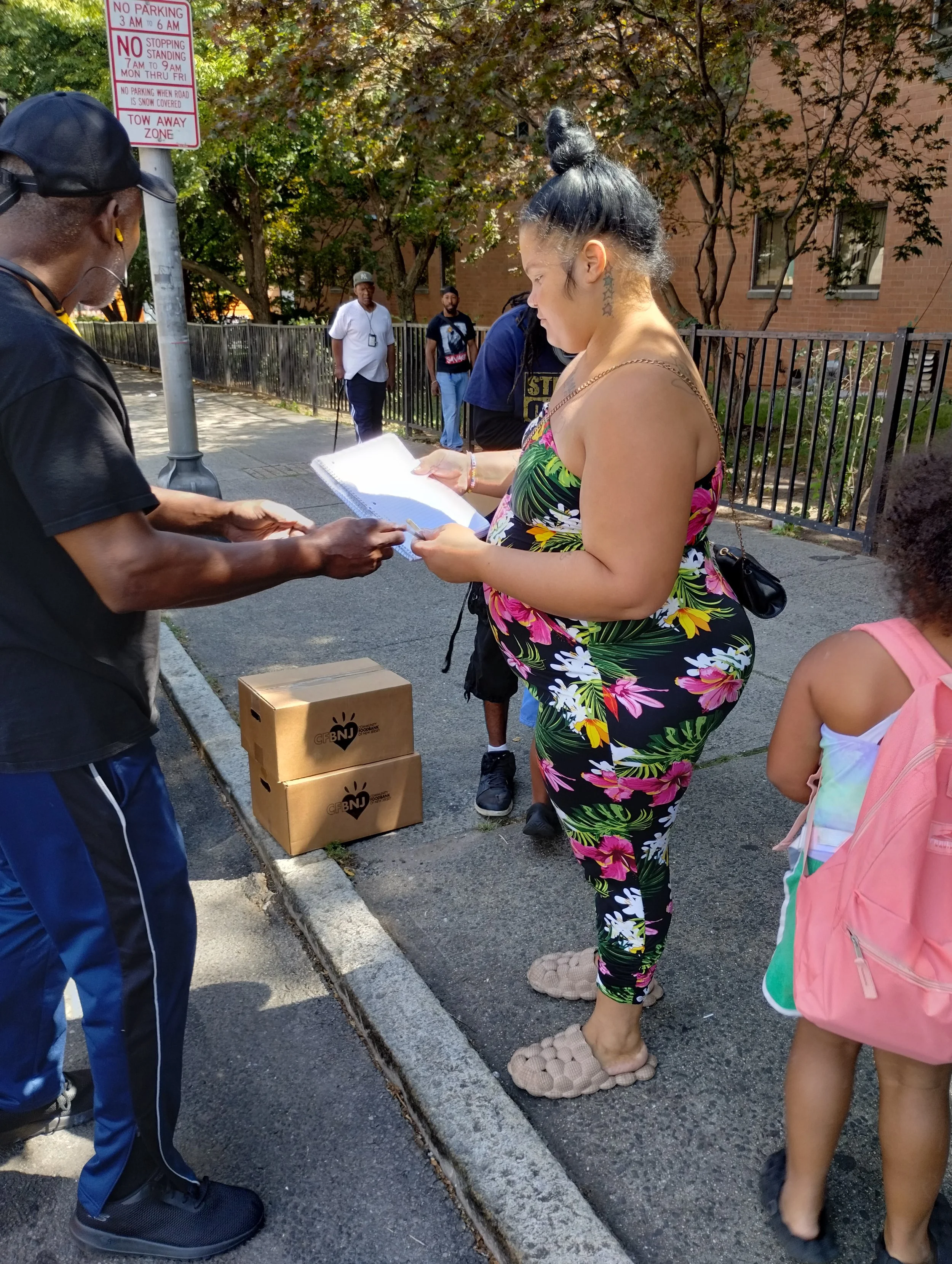 A woman in a floral jumpsuit and slippers is standing on a sidewalk, receiving a package and some papers from a man wearing a black cap and T-shirt. There are several people in the background, including a girl with a pink backpack and a man with a white shirt. There are cardboard boxes on the ground nearby, and a signpost with parking restrictions is visible. The setting appears to be outdoors in an urban area, with trees and a brick building in the background.