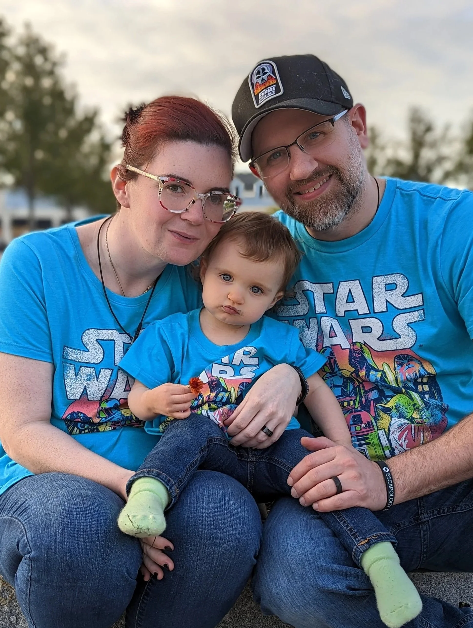 Family of three in matching blue Star Wars shirts sitting outside, with trees and a house in the background.