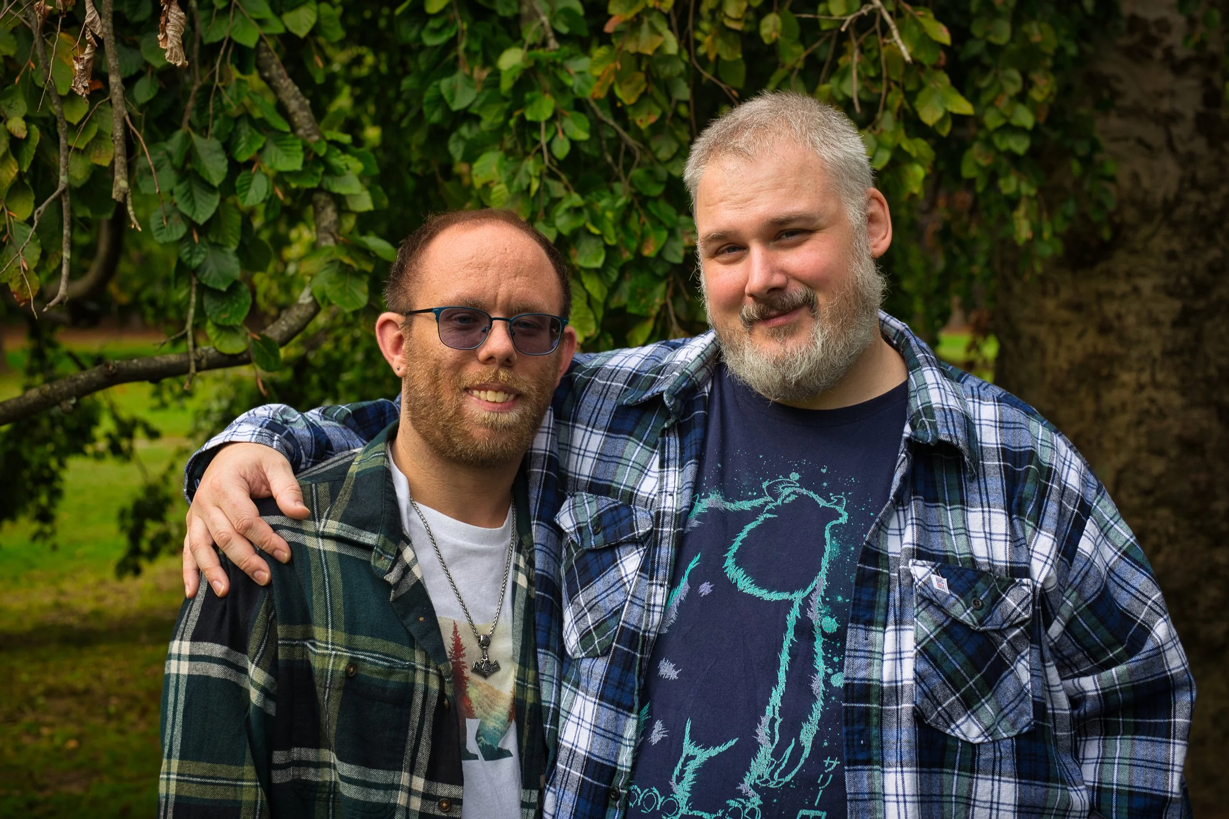 Two men standing outdoors near a tree, with one man having his arm around the other's shoulder, both smiling at the camera.