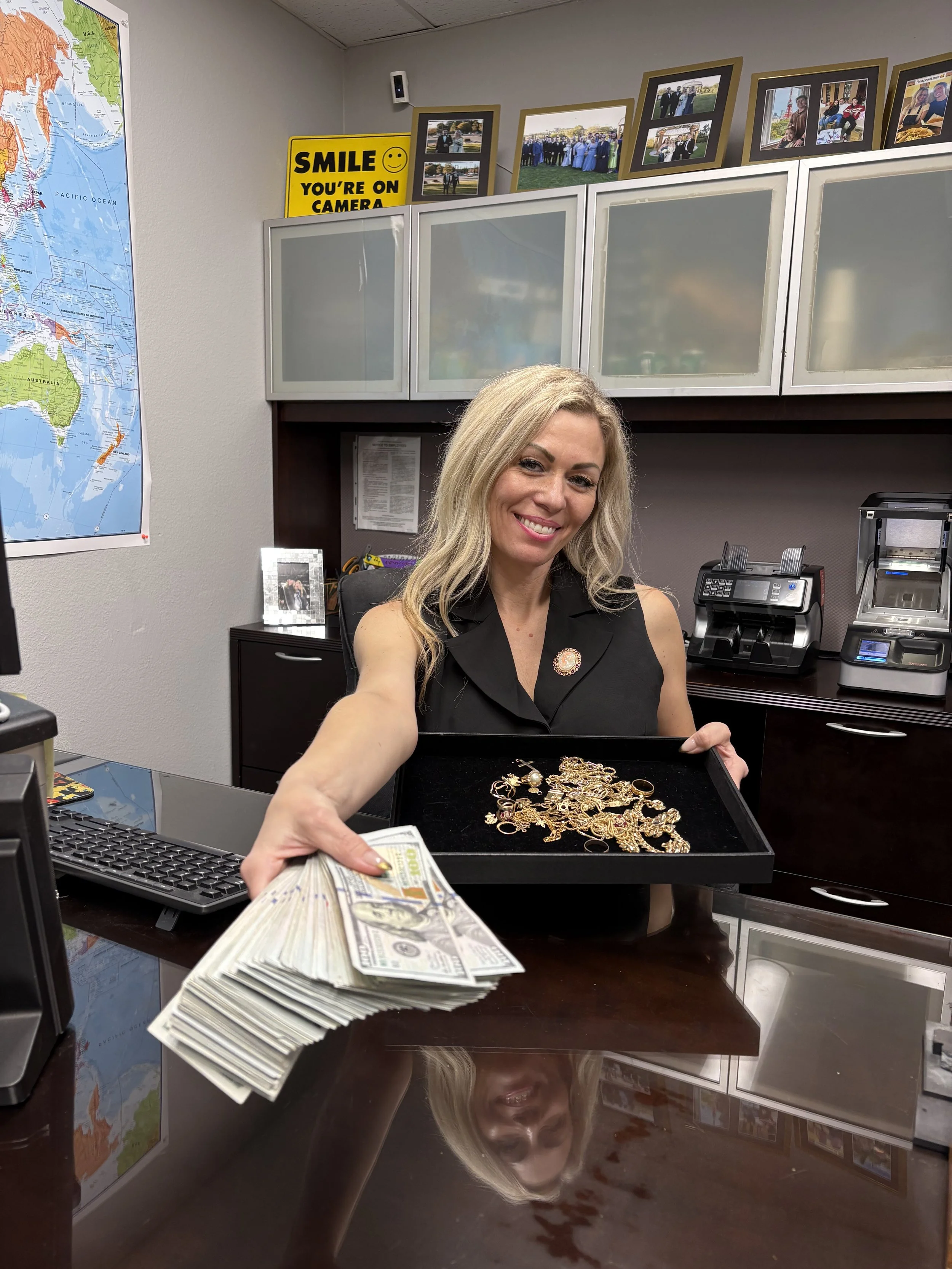 A smiling woman in a black sleeveless top is handing over a stack of hundred-dollar bills and a tray of assorted gold jewelry in an office setting.