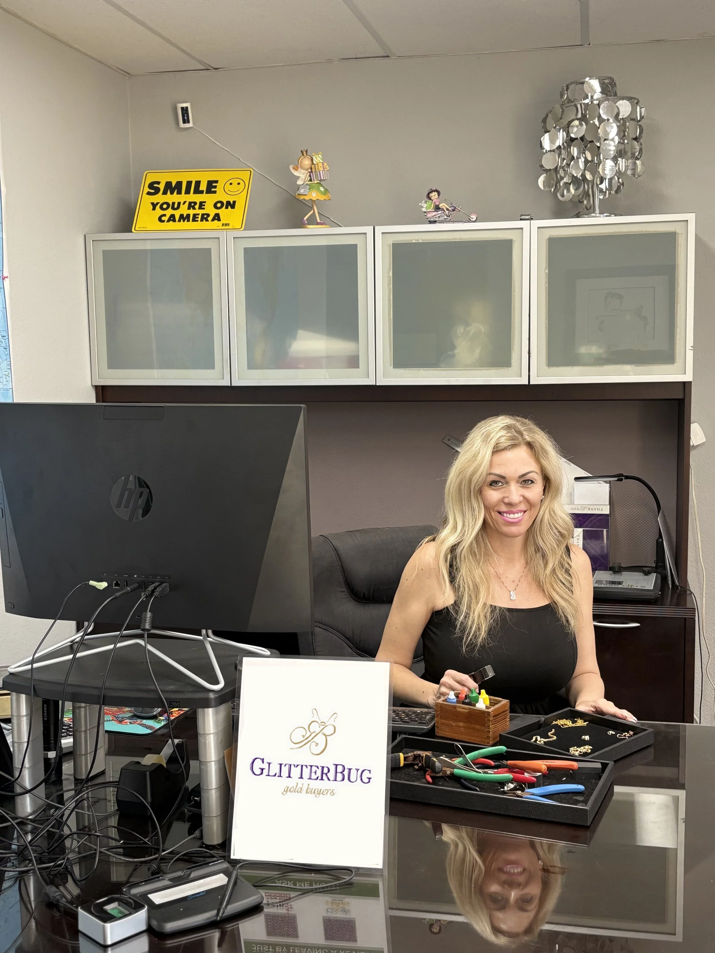 A woman sitting at a desk with jewelry supplies, a sign reading 'GlitterBug gold buyers,' and a computer monitor. Airy office space with decorative items and a 'Smile you're on camera' sign.