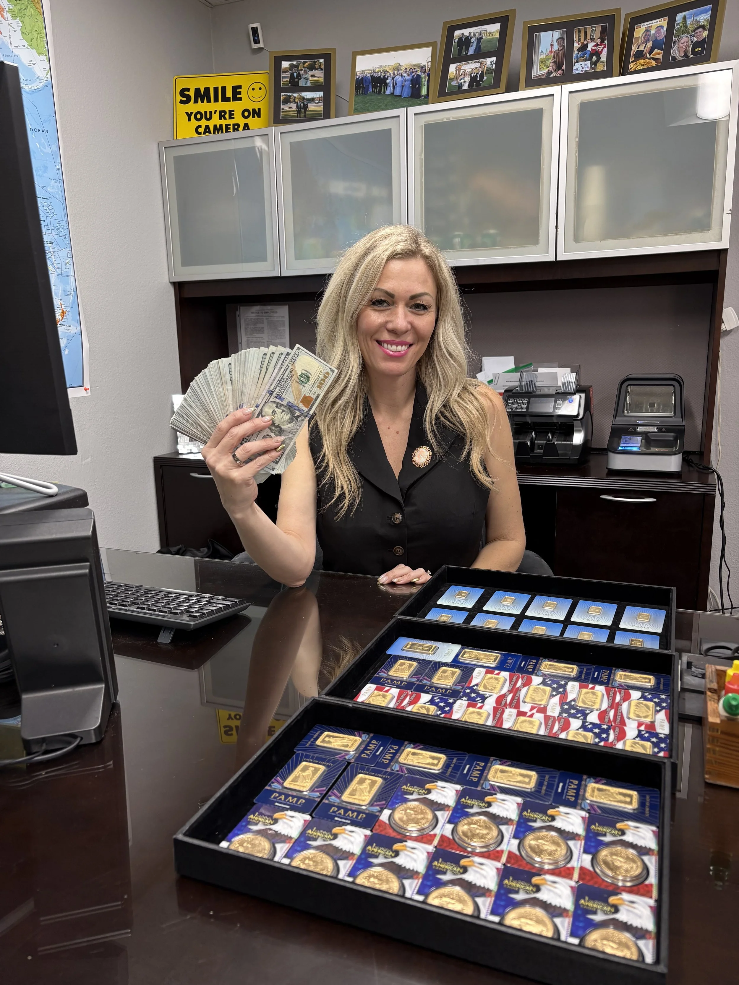 A woman with long blonde hair sitting at a desk, holding and fanning out a large amount of cash. She is smiling and wearing a sleeveless black top. On the desk in front of her are display boxes filled with gold coins and collectible cards. The background includes framed photographs on the wall, a yellow sign that reads 'Smile, you're on camera,' and office equipment like a printer and a scanner.