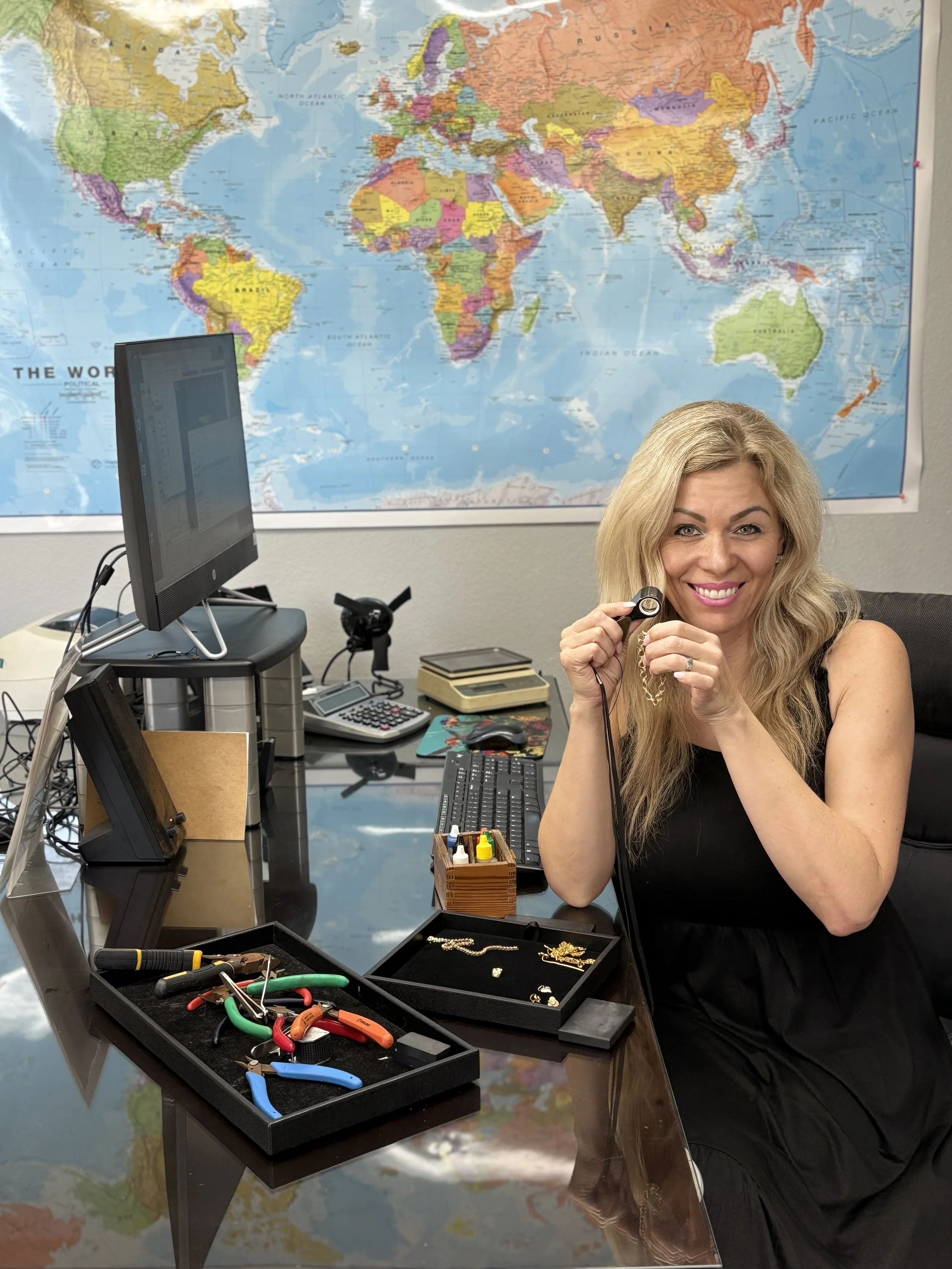 A woman with long blonde hair smiling and holding jewelry in an office, with a world map on the wall, jewelry tools, and computer equipment on her desk.