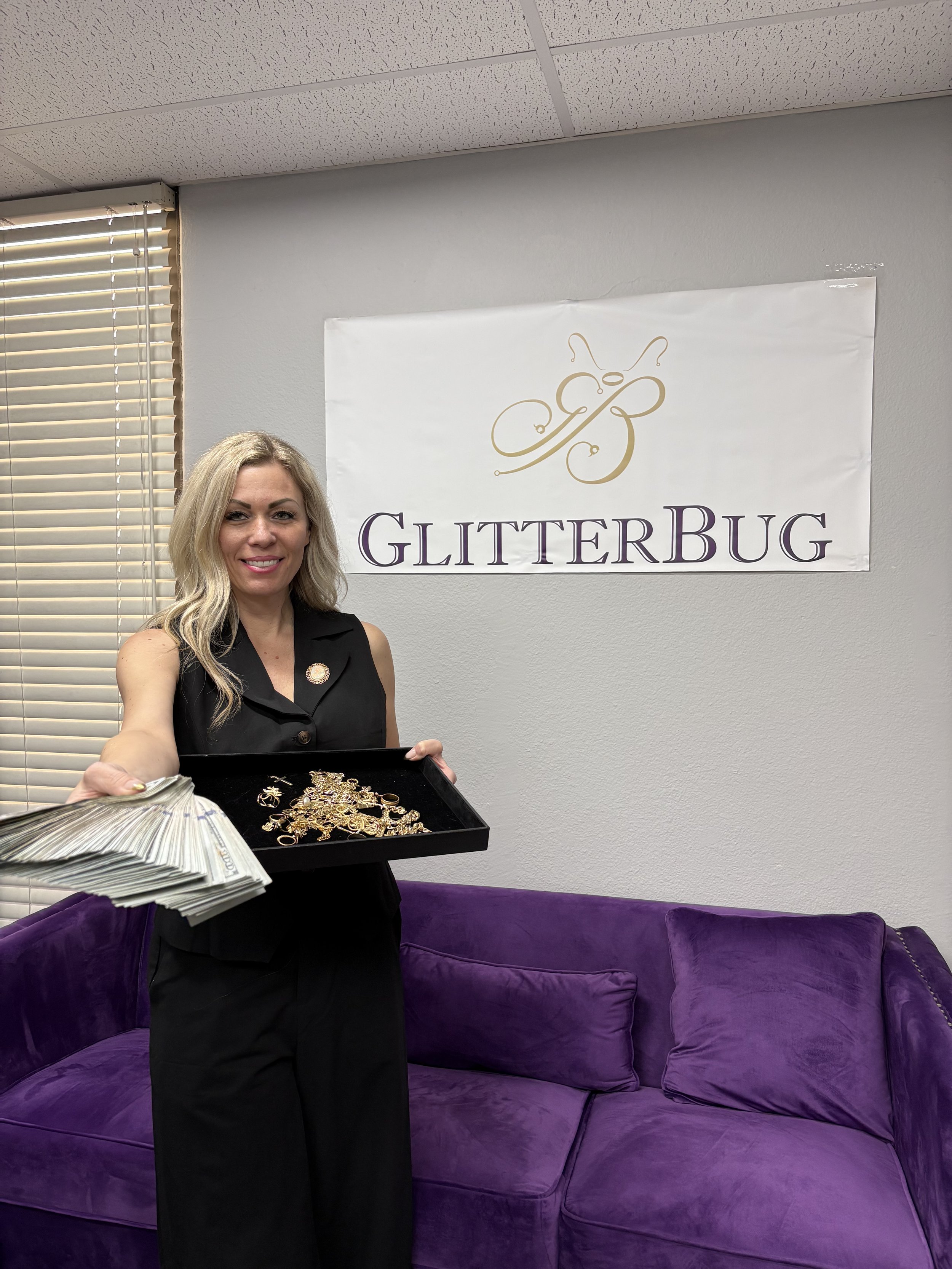 A woman with blonde hair wearing black attire is holding a tray with gold jewelry. Behind her is a purple sofa and a white wall with a glitterbug logo and text reading "GlitterBug."