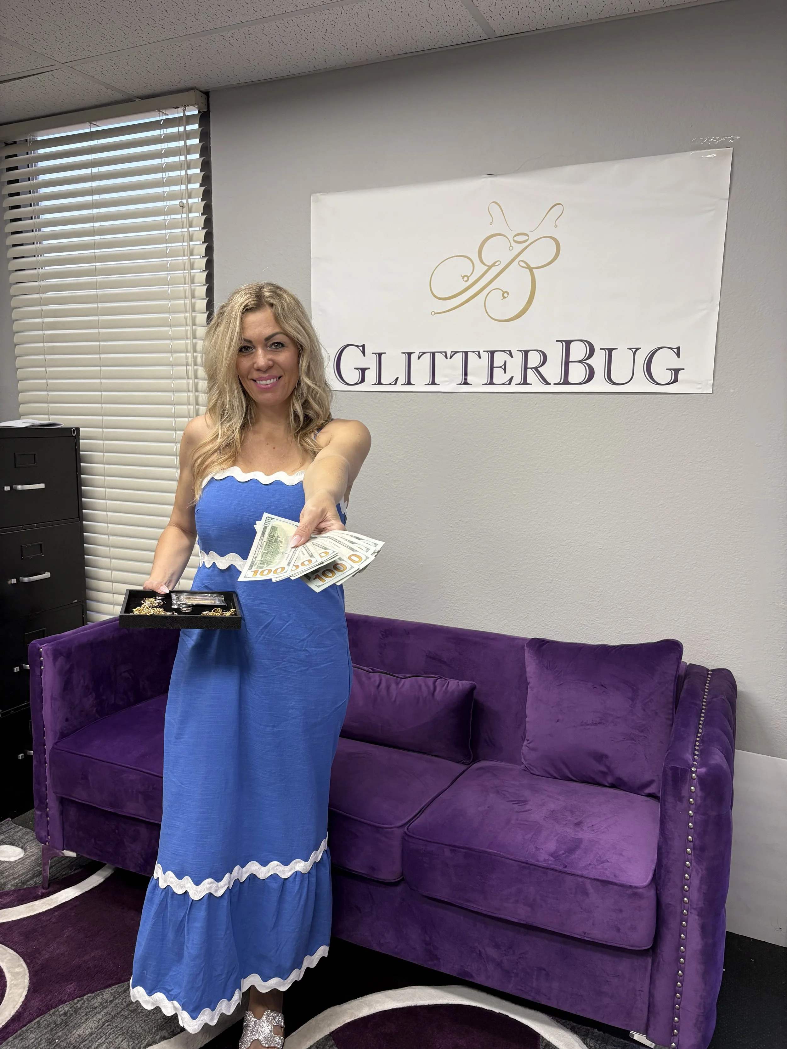 A woman in a blue dress holding dollar bills and jewelry, standing next to a purple sofa in an office with a 'GLITTERBUG' sign on the wall.