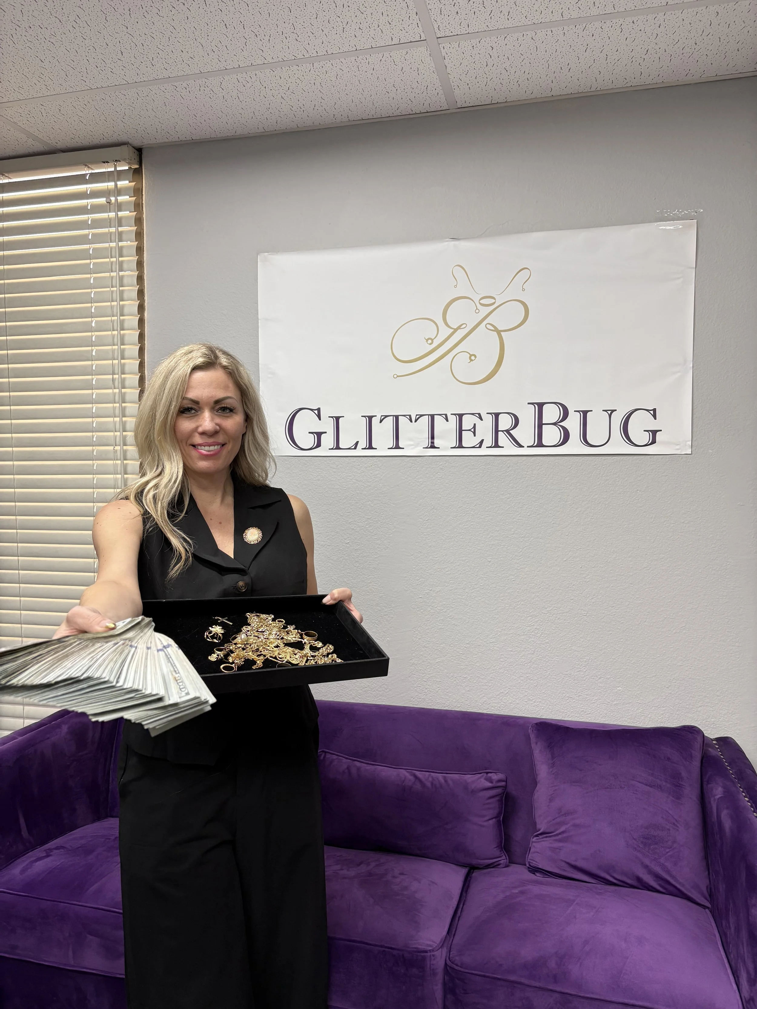 A smiling woman holding a tray filled with jewelry, standing in front of a banner that reads 'GlitterBug' with a butterfly logo, inside a room with a purple sofa and blinds on the window.