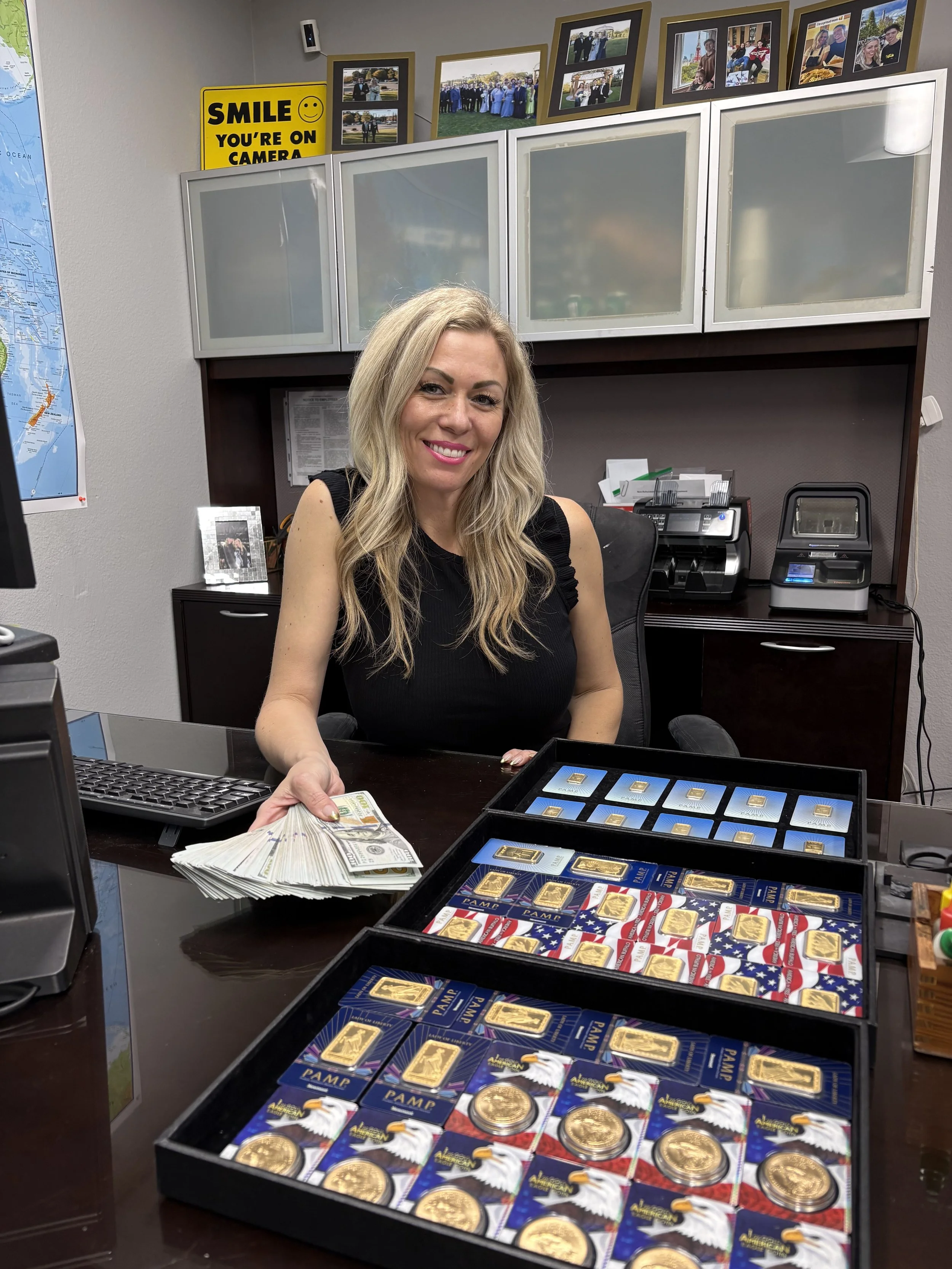 A woman sitting at a desk in an office displaying a collection of gold and silver commemorative coins and bars, with cash in her hand, smiling at the camera.
