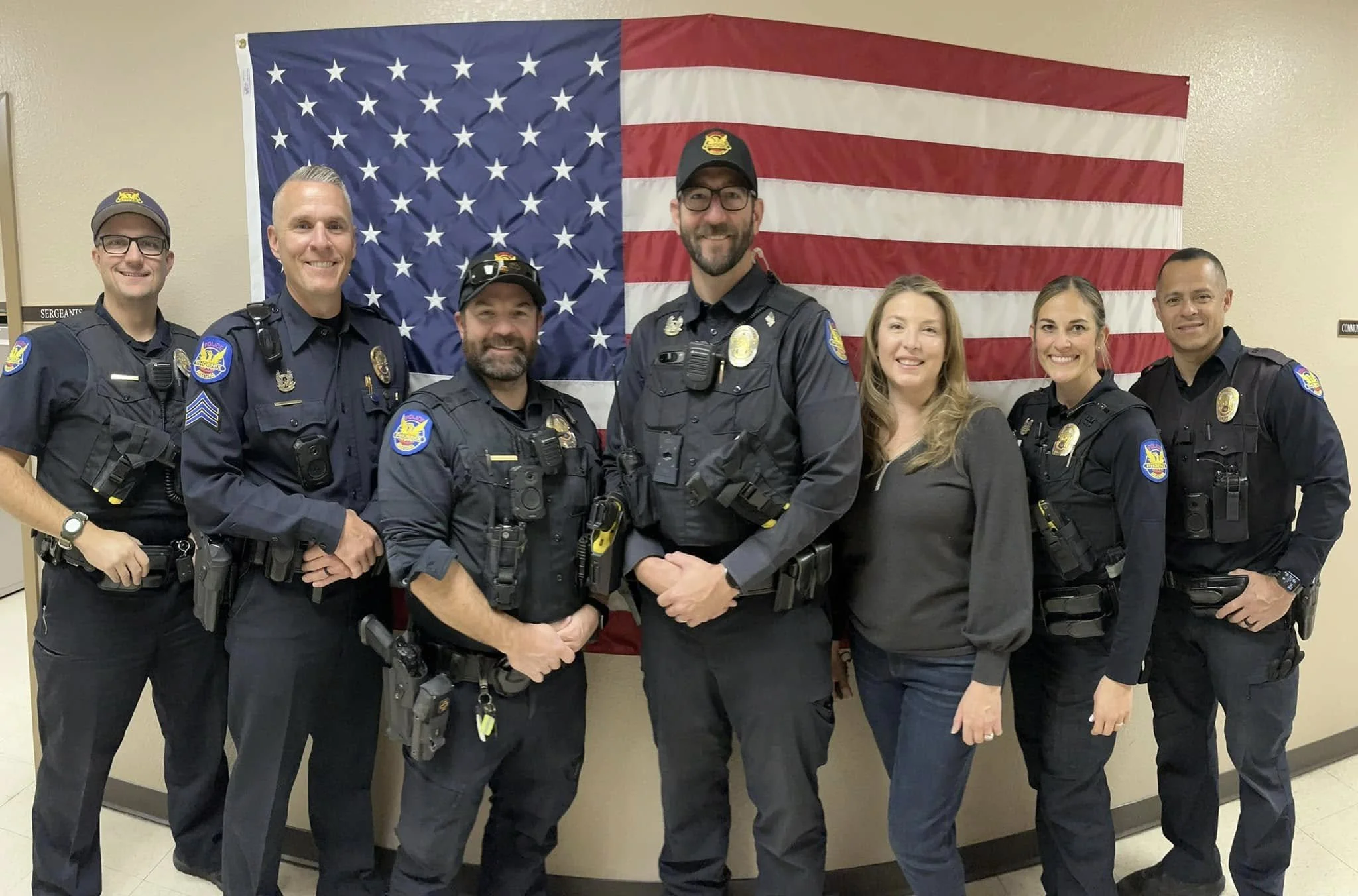 Seven police officers and one civilian woman standing in front of an American flag. The officers are in uniform, some wearing caps, and smiling at the camera.
