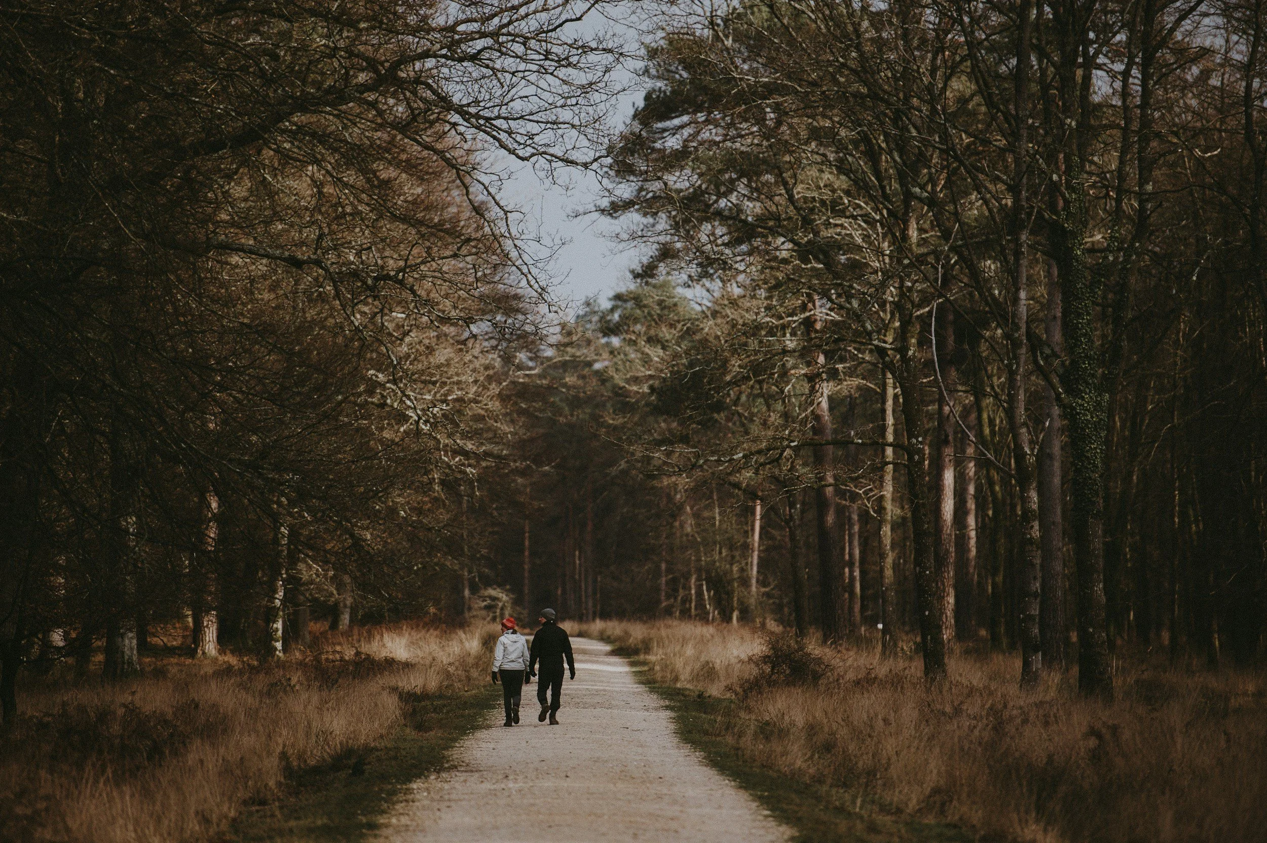 A man and woman walking on a dirt path through a forest with tall trees and dry grass