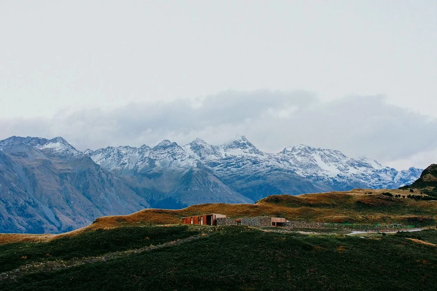 A scenic landscape with snow-capped mountains in the background, rolling grassy hills in the middle ground, and a modern building on the hillside in the foreground.