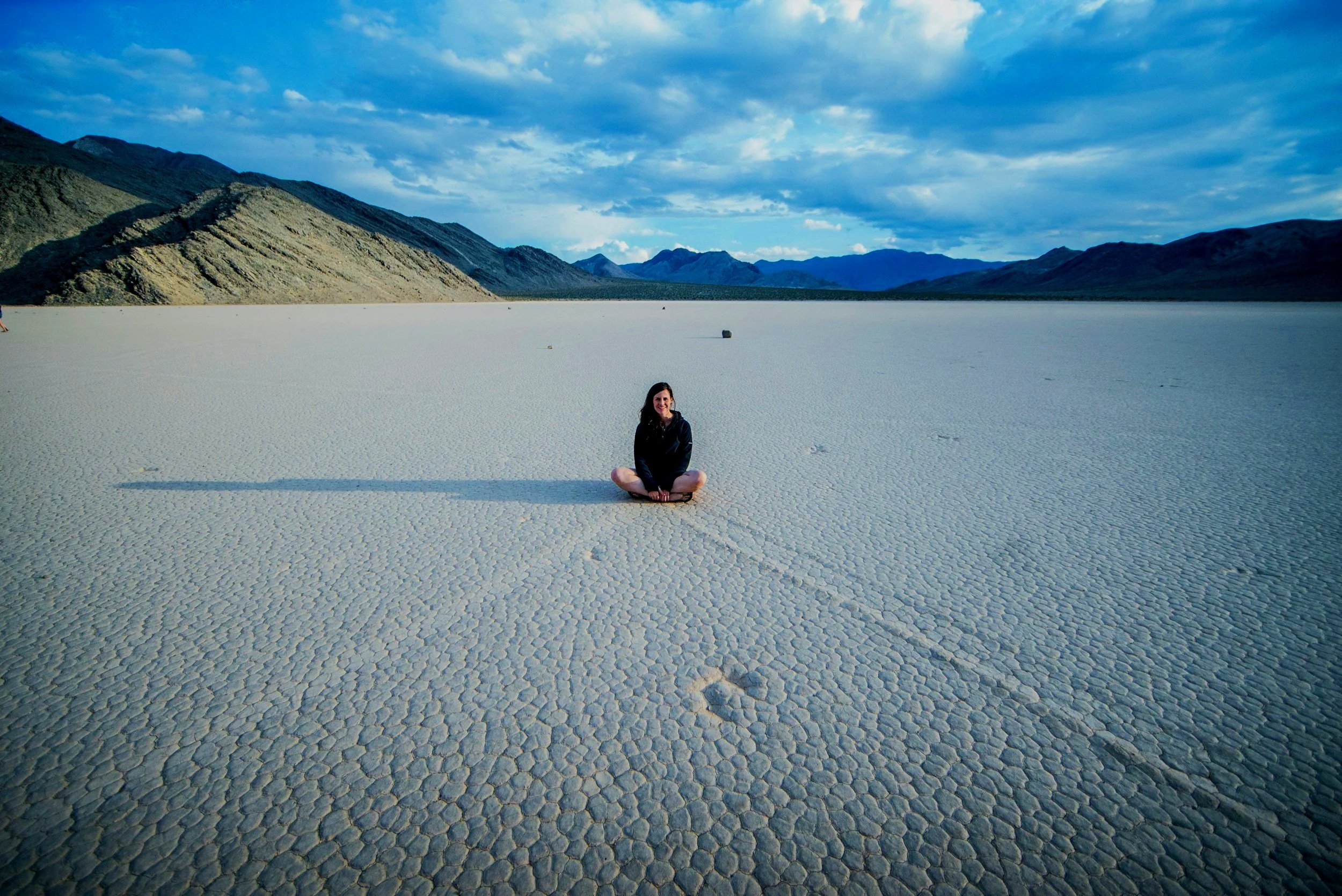A woman sitting cross-legged on a vast, cracked salt flat with mountains in the background under a partly cloudy sky.