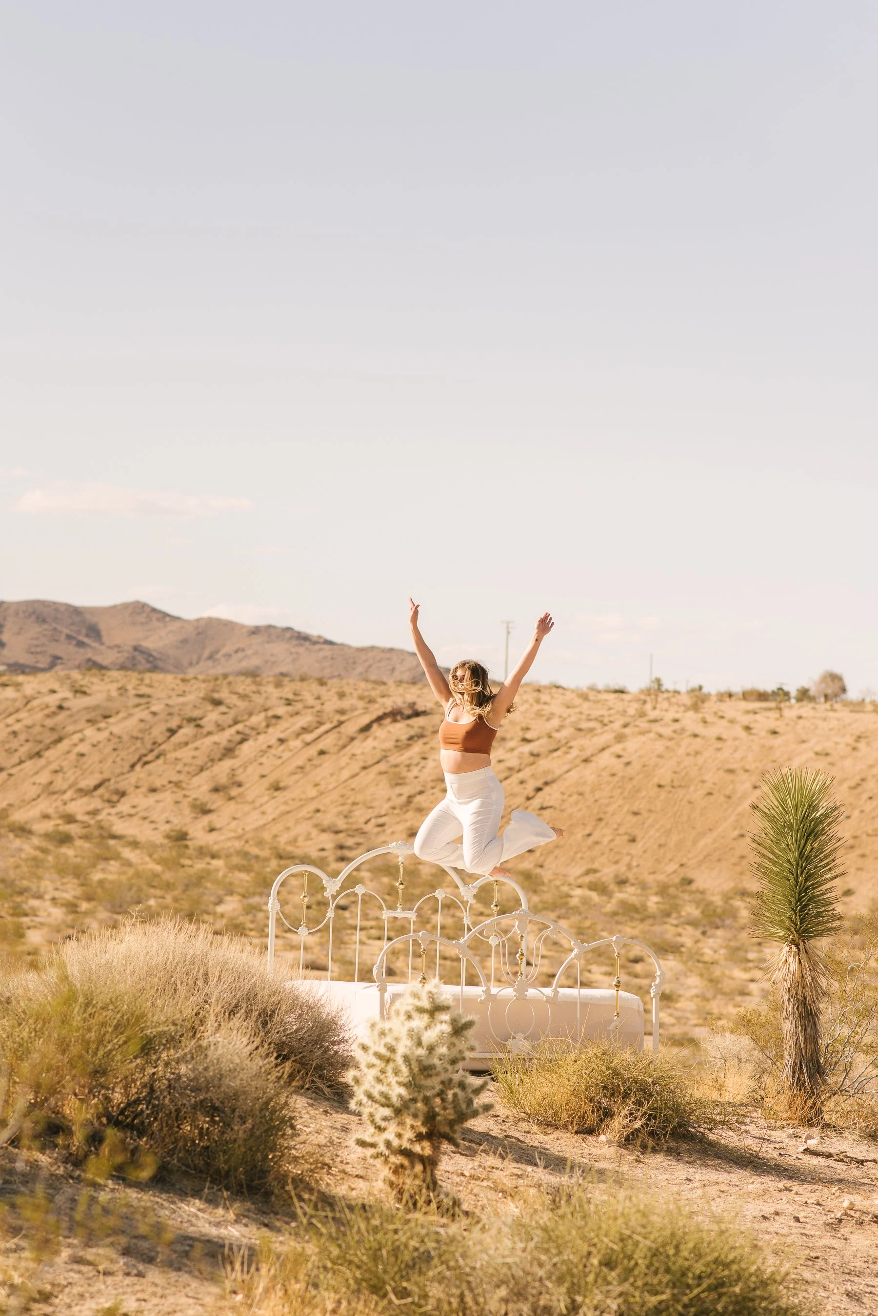 A woman jumping on a white metal bed frame in a desert landscape with shrubs and mountains in the background.