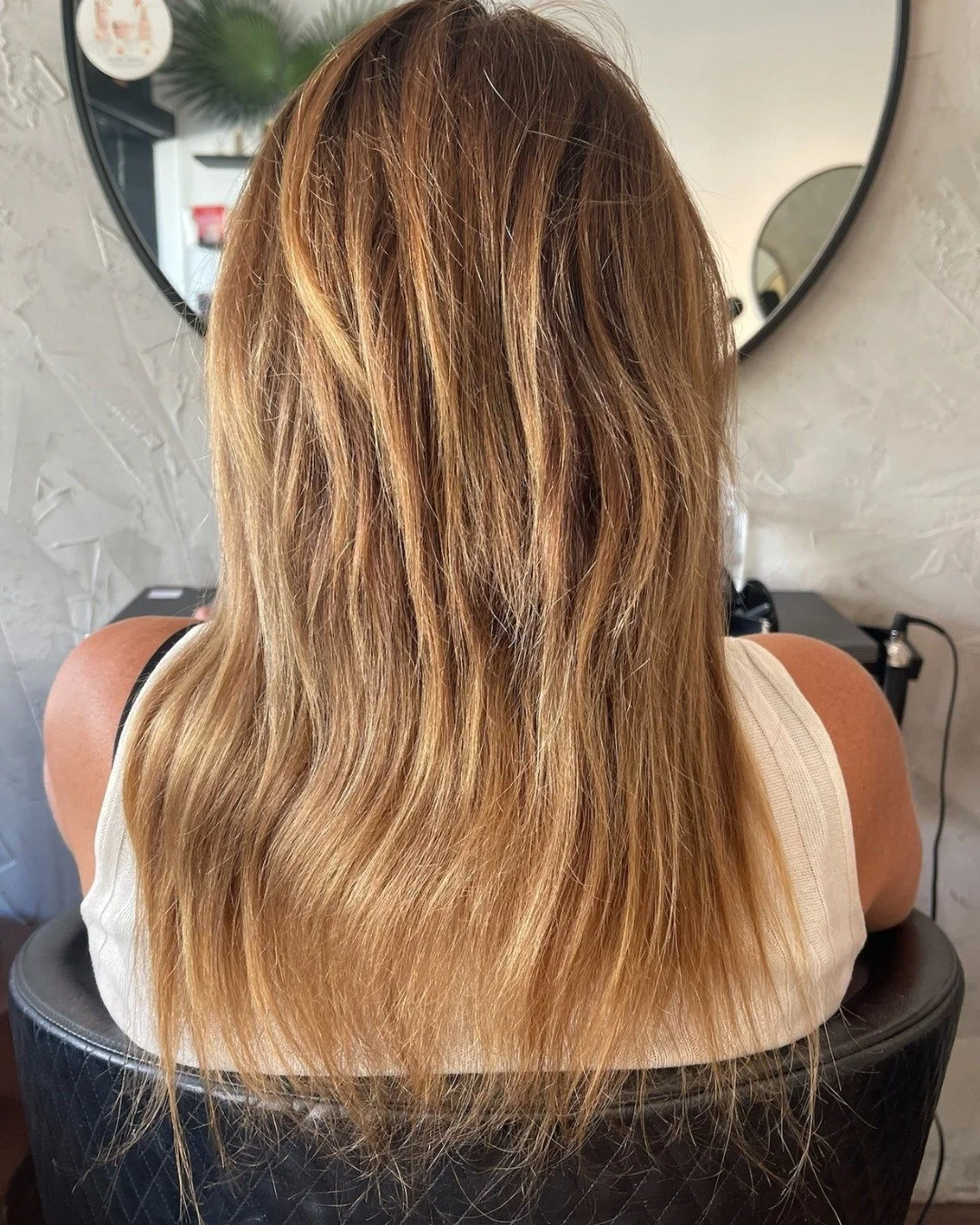 Back of a woman with long, wavy, blonde hair sitting in a salon chair in front of a mirror.