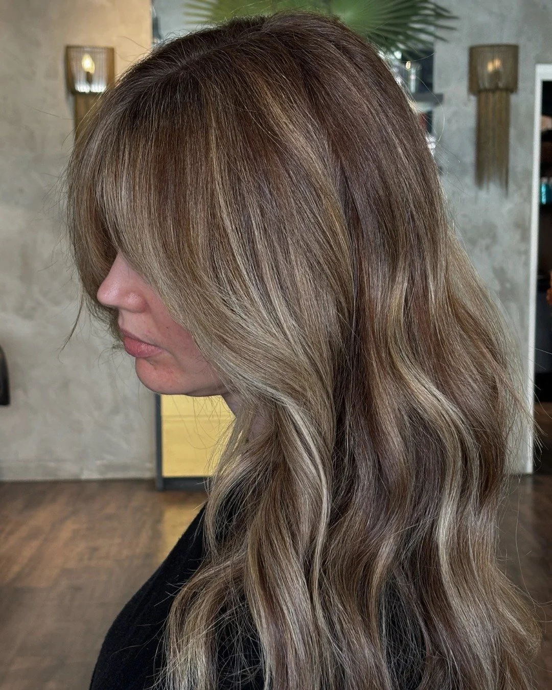 Side view of a woman with long, wavy hair in shades of blonde and light brown, standing indoors with a wall-mounted light fixture and a plant in the background.