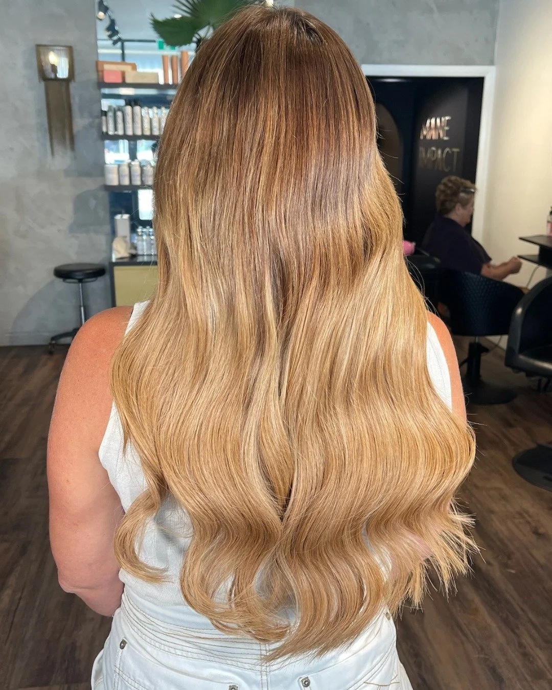 Back view of a woman with long, wavy blonde hair in a salon. There is a woman seated in the background, and salon shelves with hair products are visible.