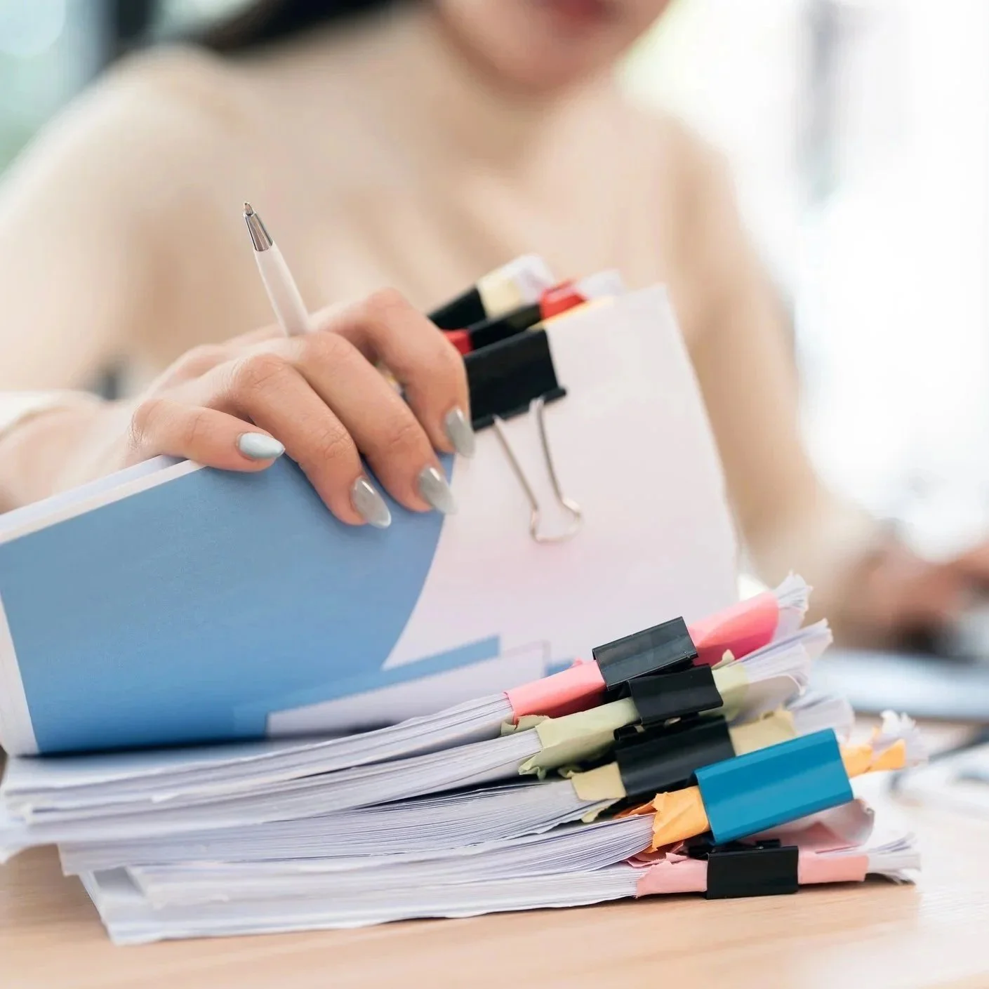 Hand holding a pen and organizing a large stack of papers clipped with multiple colorful binder clips and sticky notes.