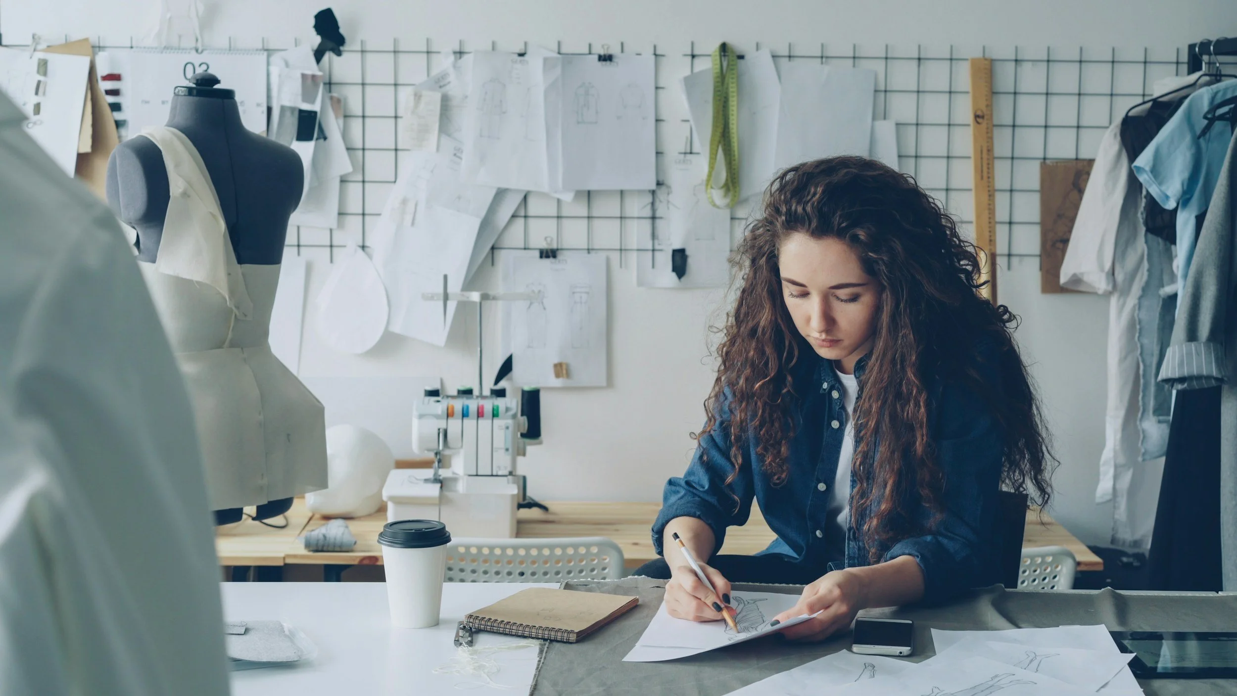 Young woman working on fashion designs at her workspace in a fashion studio, surrounded by dress patterns, sketches, a mannequin, and measuring tools.