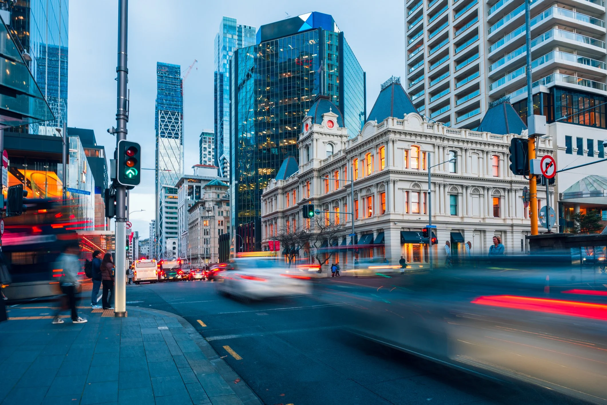 Auckland City street scene in downtown with modern glass skyscrapers and historic white building, busy with cars and pedestrians at dusk.