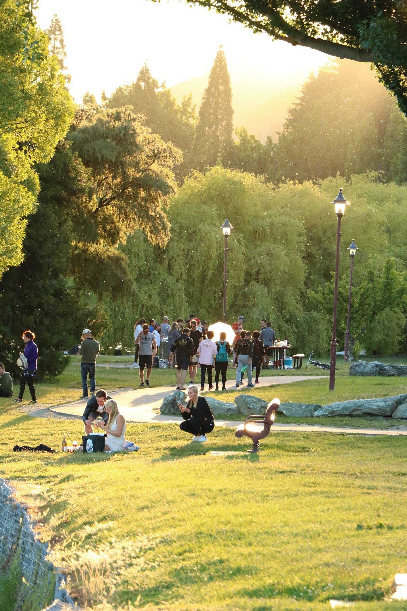 People enjoying a gathering in a lush park during sunset, with trees, lampposts, and large rocks in the background.