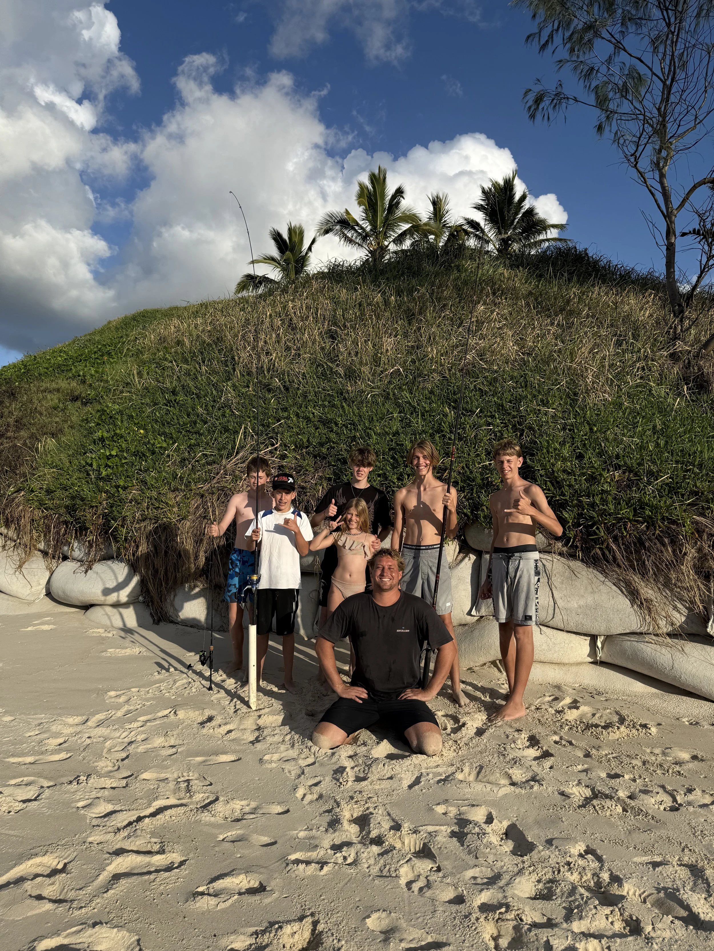 Group of six children and one adult on a sandy beach holding fishing rods, with a grassy hill, palm trees, and a partly cloudy sky in the background.