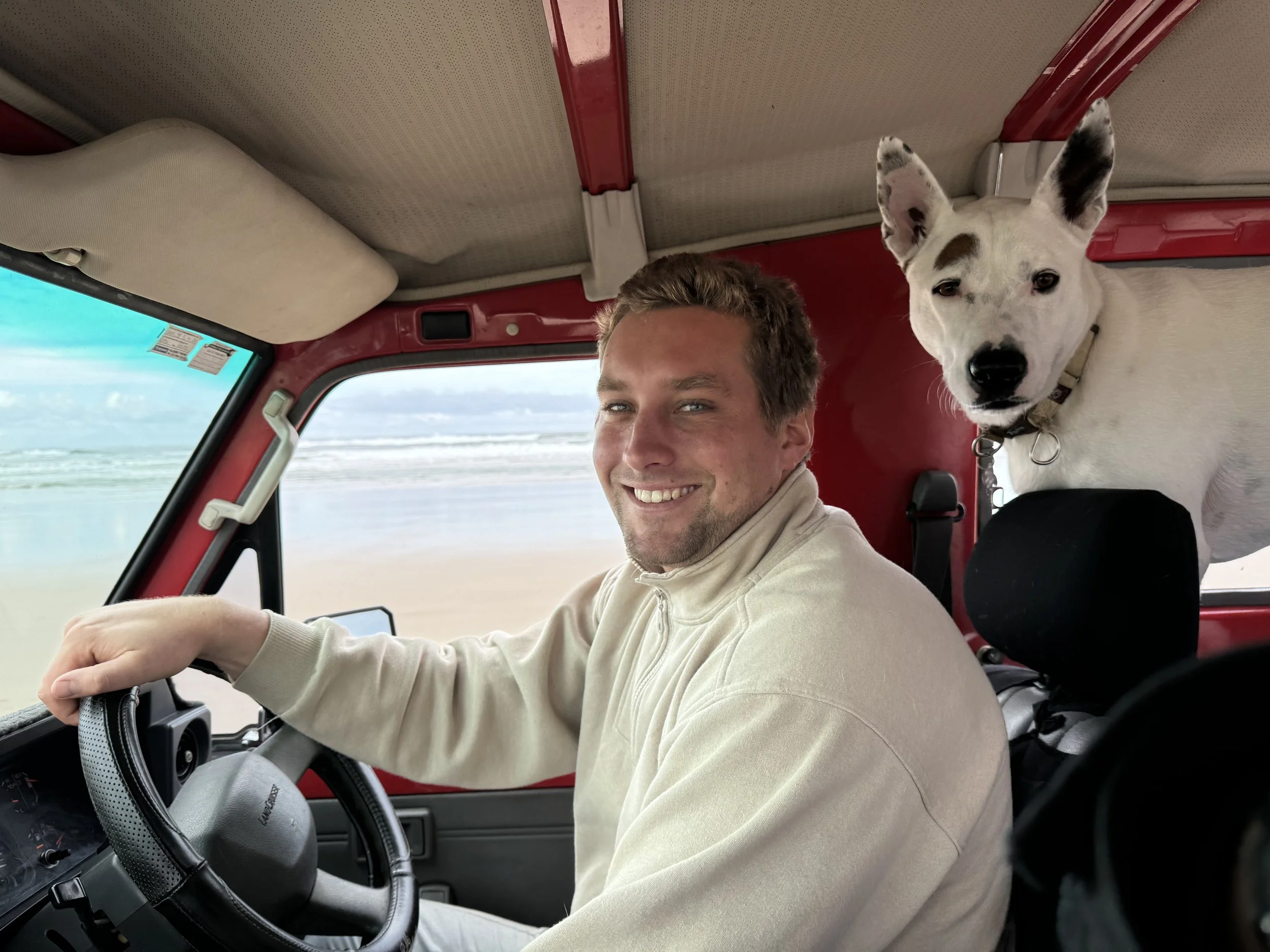 A smiling man driving a vehicle on the beach, with a white dog sitting behind him.
