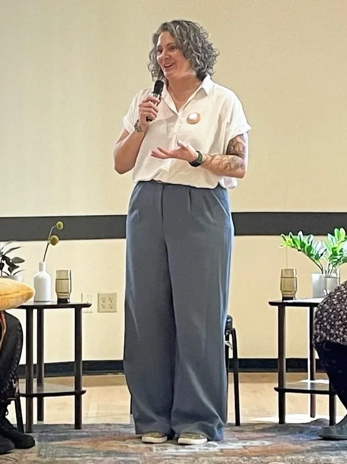 Woman with curly, shoulder-length hair speaking into a microphone wearing a white shirt and blue pants
