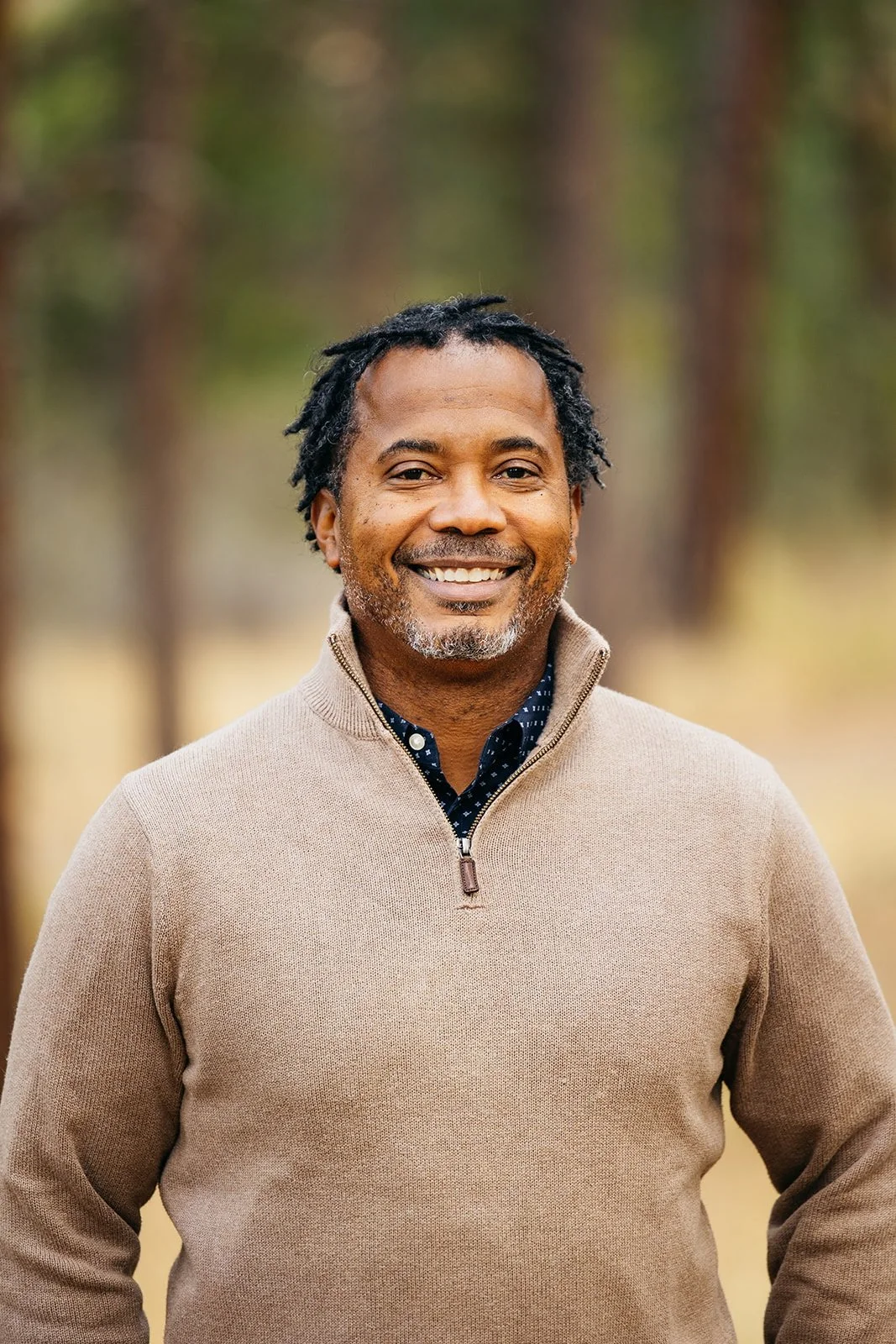 A middle-aged man with black dreadlocks and a beard, smiling, wearing a beige zip-up sweater over a dark blue shirt with white polka dots, standing outdoors with trees in the background.