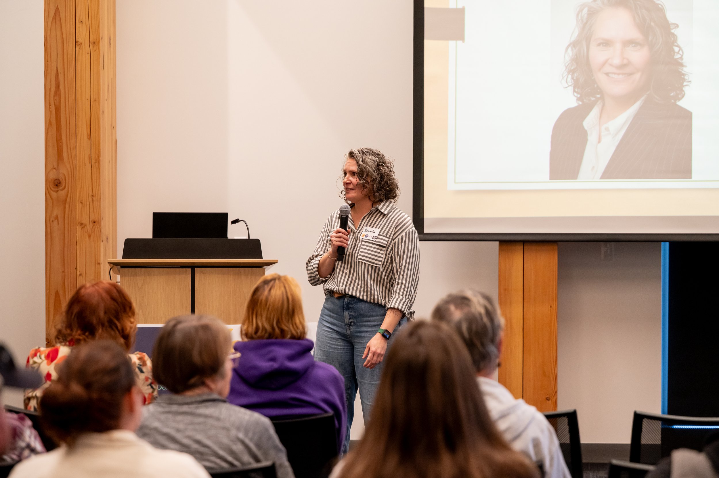 A woman with curly hair holding a microphone presents in front of an audience at a conference room, with a large screen showing her portrait on the wall behind her.