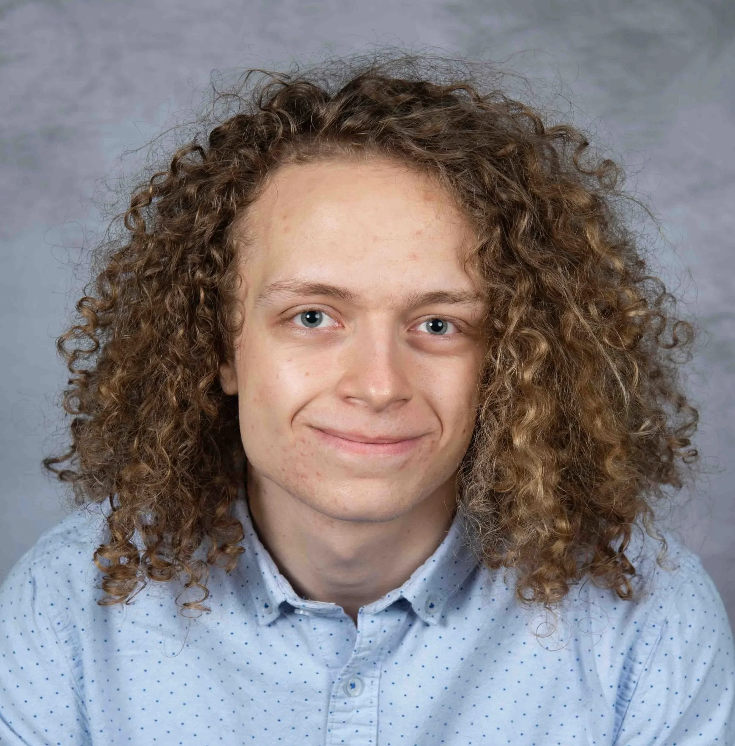 Smiling white male with longer, curly light brown hair, blue eyes wearing a blue shirt