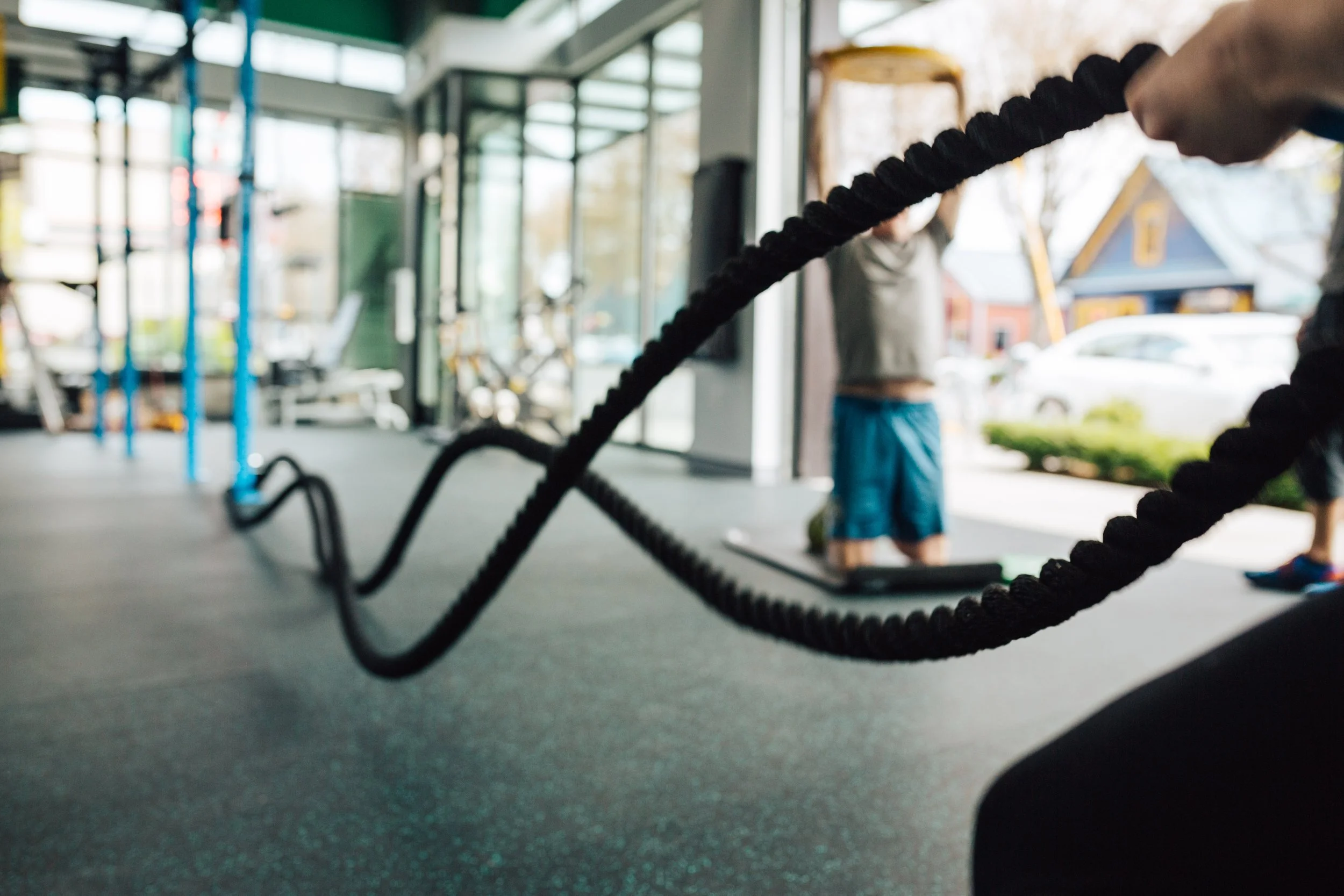 Close-up of black battle ropes in focus at a gym, with a person in workout attire blurred in the background, inside a fitness center with large windows and some outdoor scenery visible.