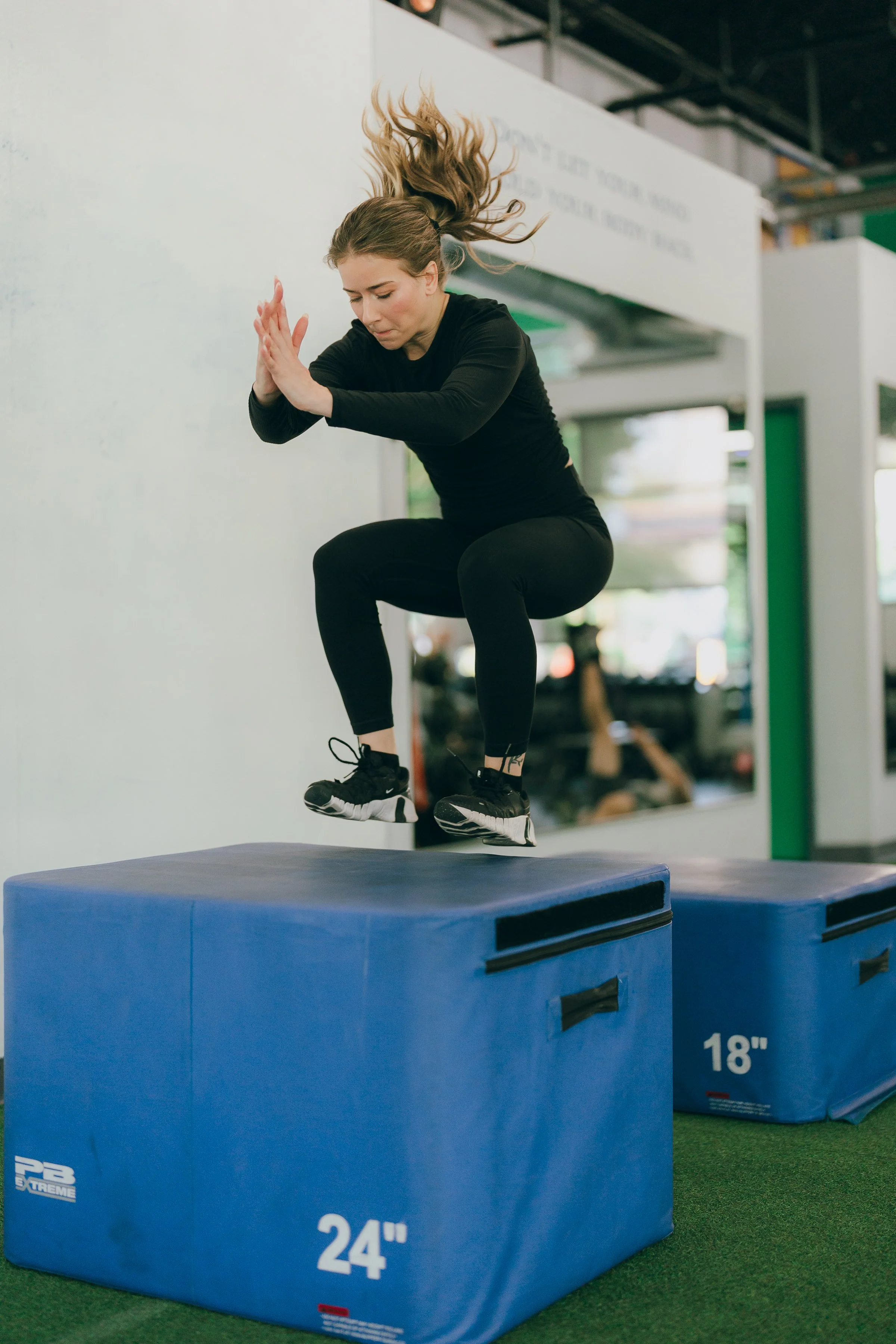 A Female Personal Trainer jumping onto a blue exercise box at a gym in The fremont neighborhood of seattle, wearing black athletic clothing and sneakers.